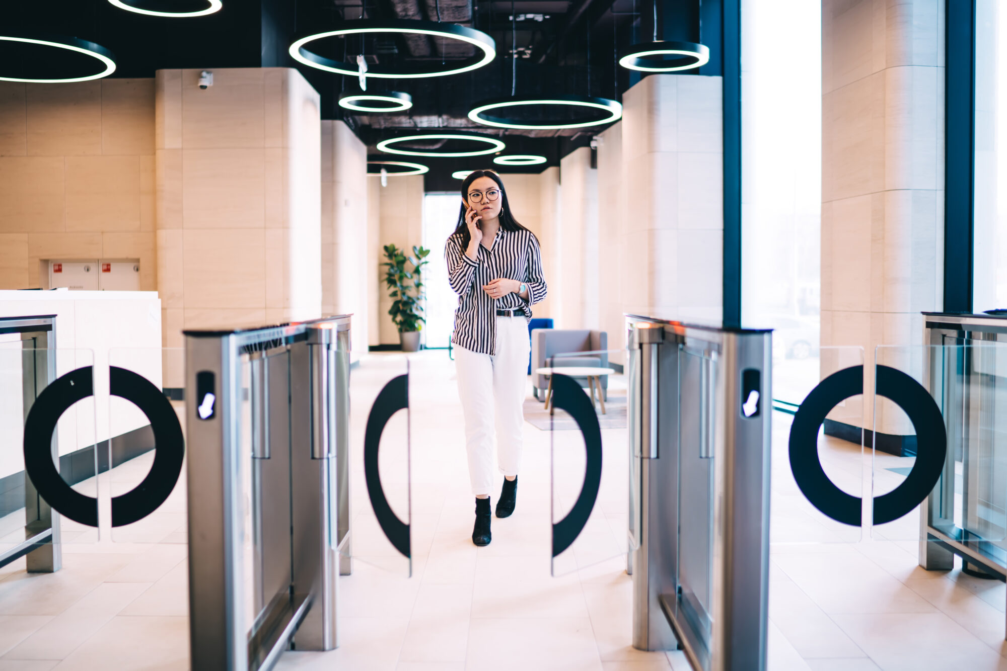 A woman in a striped shirt and white pants walks through modern electronic access control glass turnstiles in an office building. Circular ceiling lights illuminate the space, while large windows provide natural light. She's holding a phone to her ear.