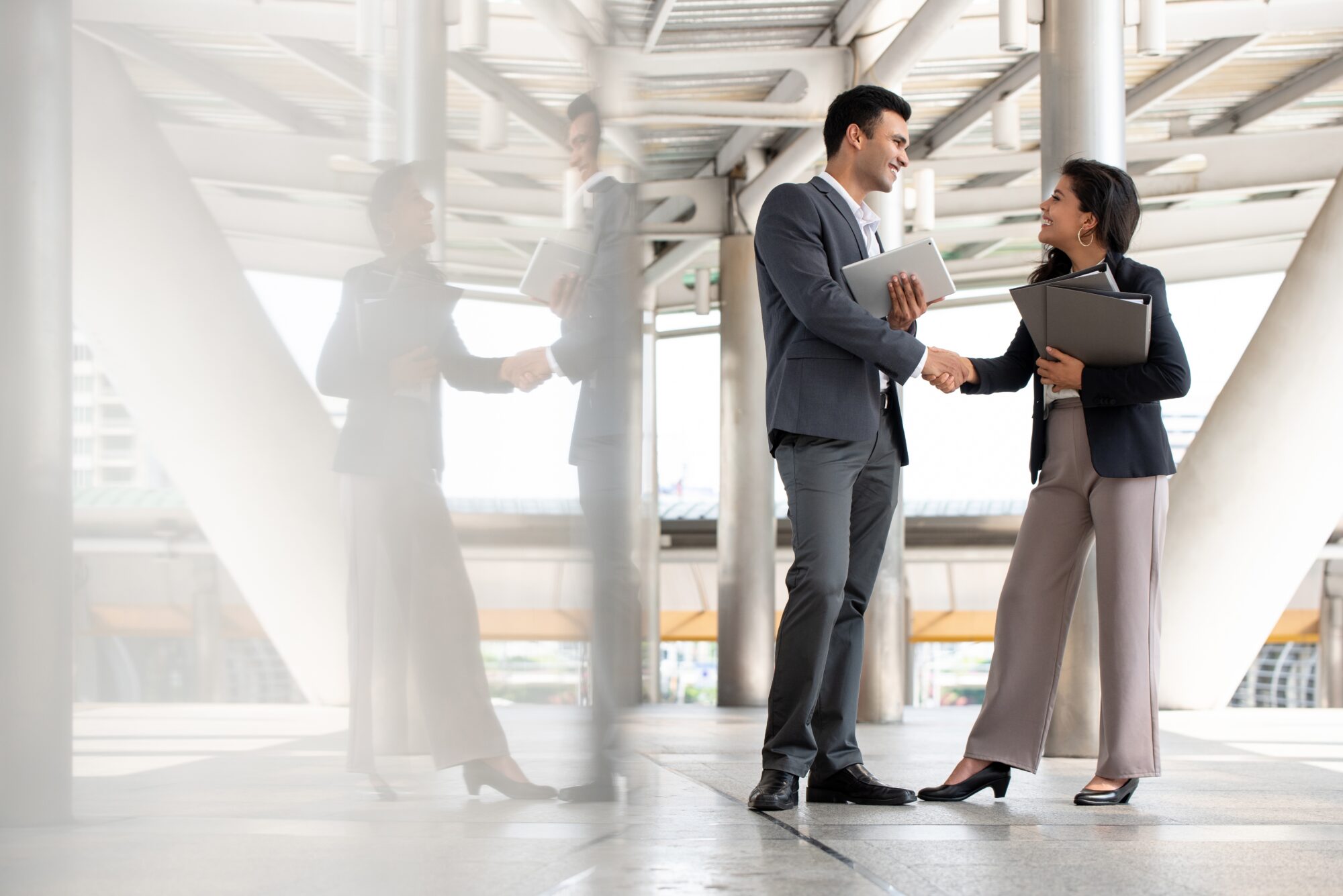 A man and woman in business attire are shaking hands outdoors, smiling as they hold documents about difficult tenants. Standing by a large glass window that reflects their images, the setting appears to be a modern architectural structure.