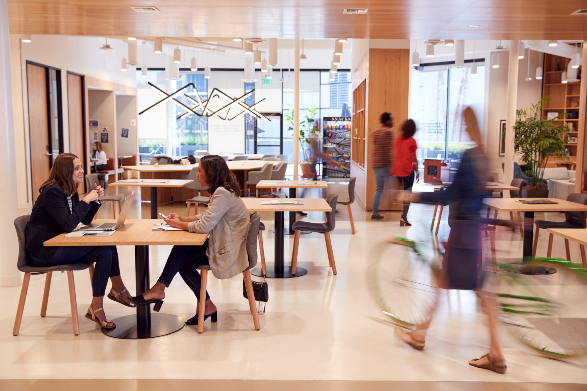 A modern office space buzzes with activity as people work and walk around. Two women sit at a table engaged in conversation, while touchless access control ensures seamless entry. A person rides a bicycle past in the foreground, all within a well-lit space accented by wood and greenery.