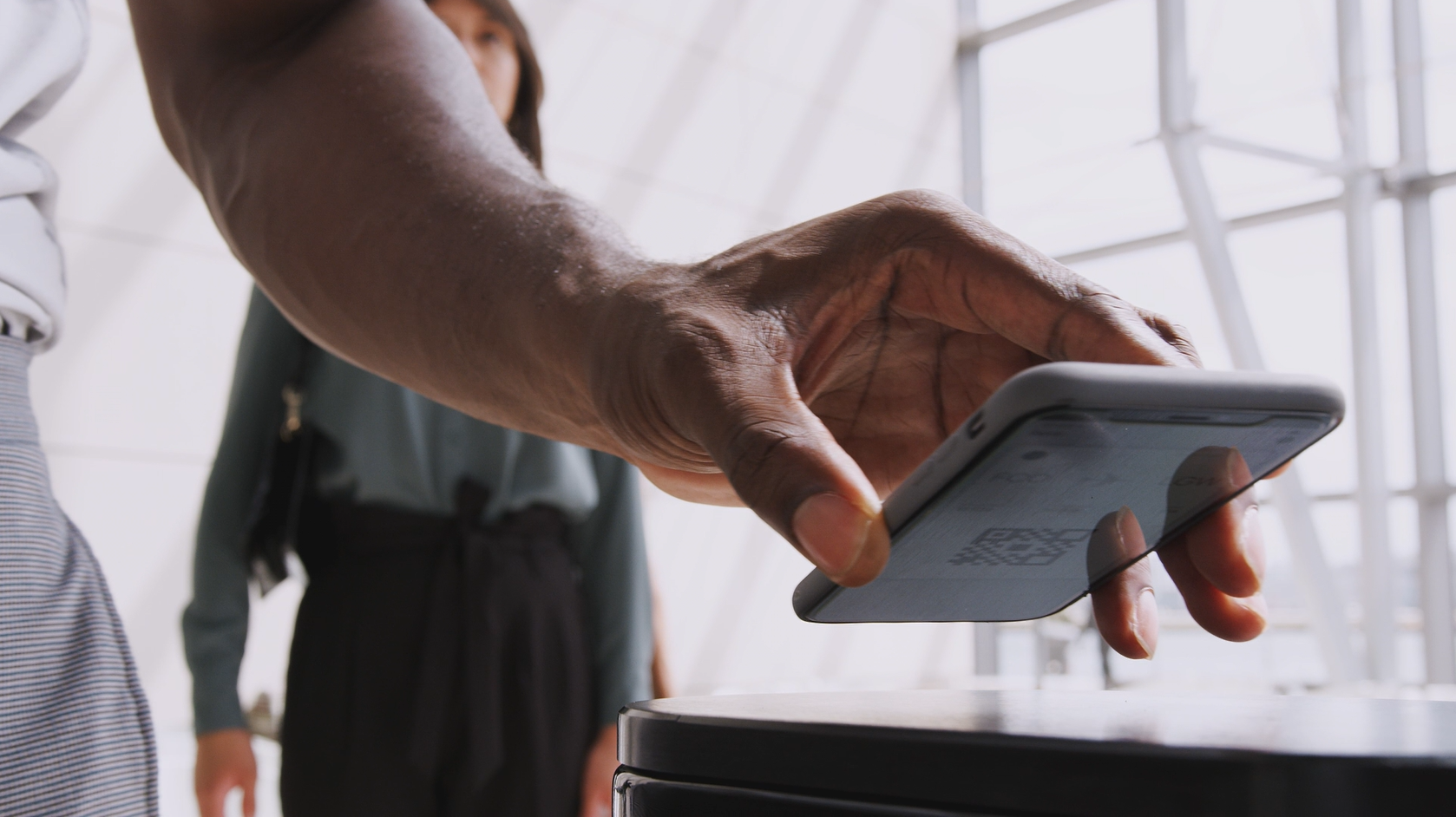 A close-up of a person using visitor management software on their smartphone, scanning a QR code at a payment terminal. In the background, another individual waits. The setting is a modern indoor space with large windows, exuding sleek sophistication.