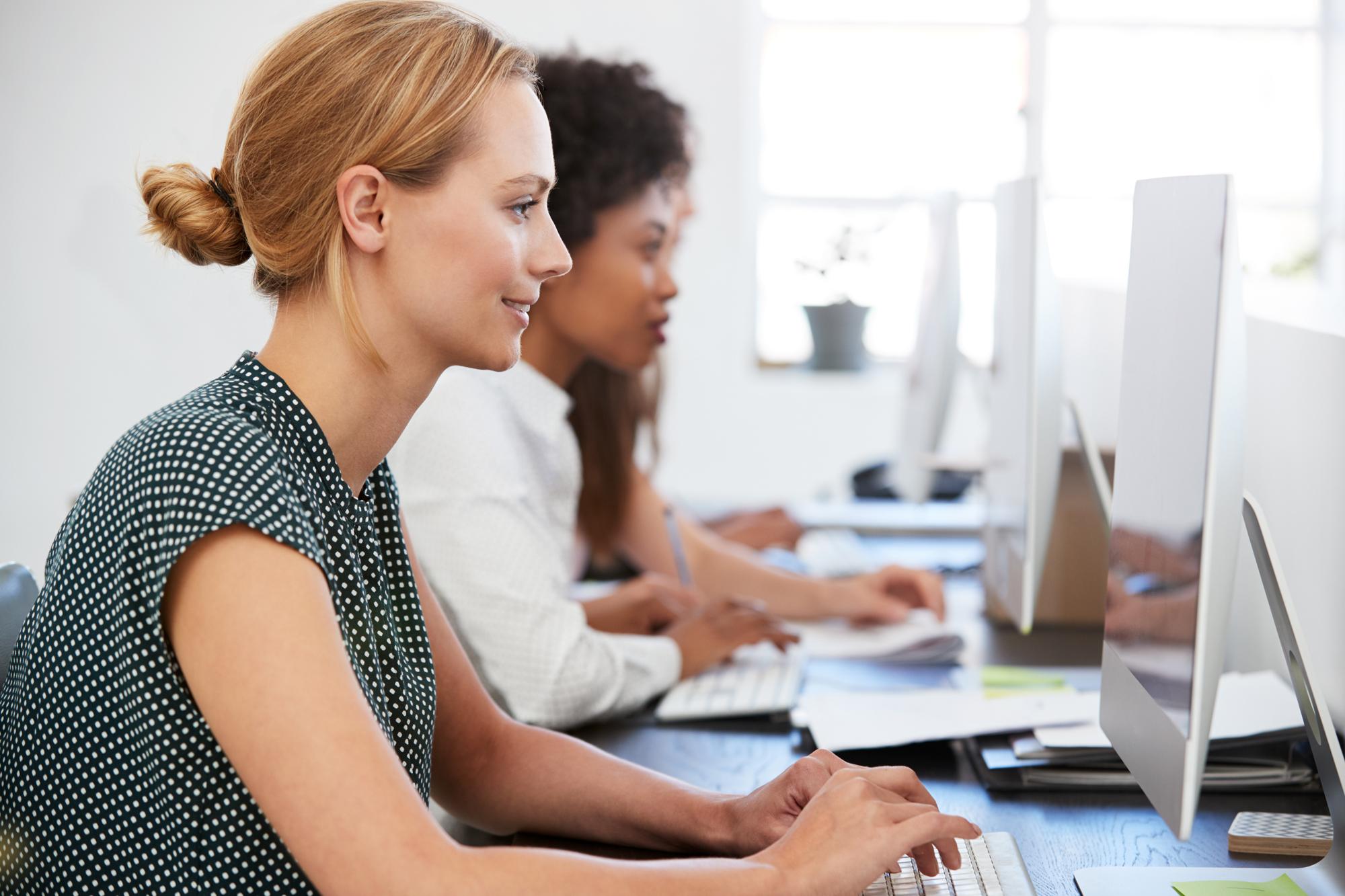 Two women are sitting side by side at desks, focused on desktop computers, engaged in a genea-related project. They are in a well-lit office space with large windows, both typing on keyboards, suggesting a collaborative or work-related activity.