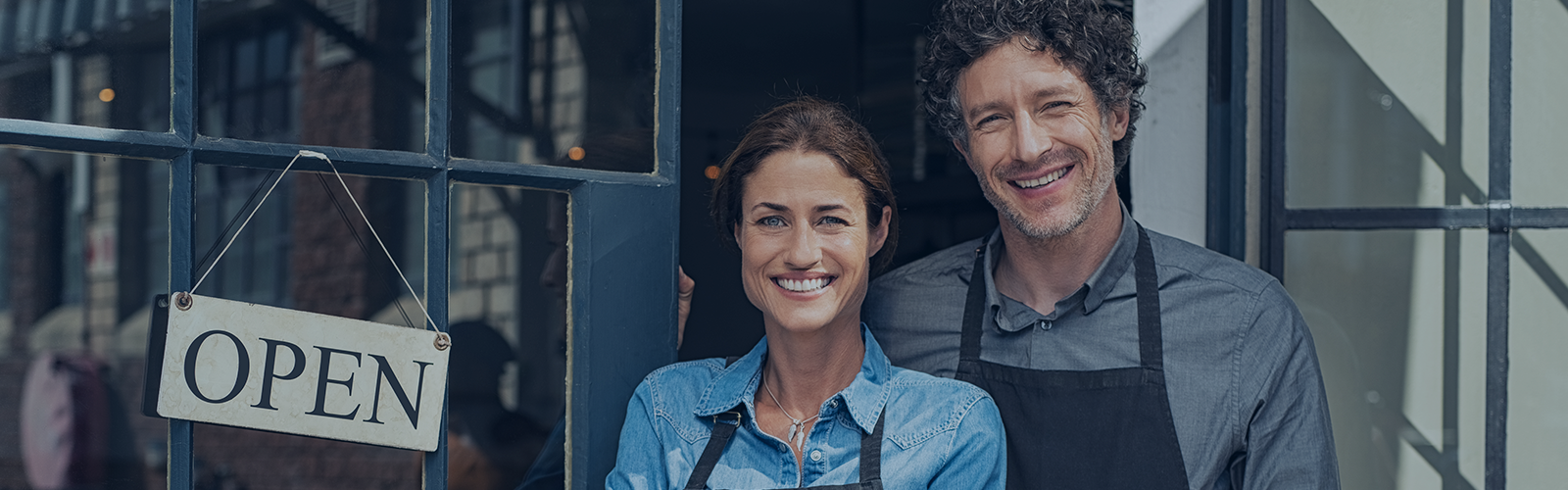 A smiling man and woman wearing aprons stand in the doorway of a shop with an OPEN sign, embodying the essence of success. The background reveals glass windows, offering a glimpse of their well-managed space, enhanced by effective access control for small business operations.