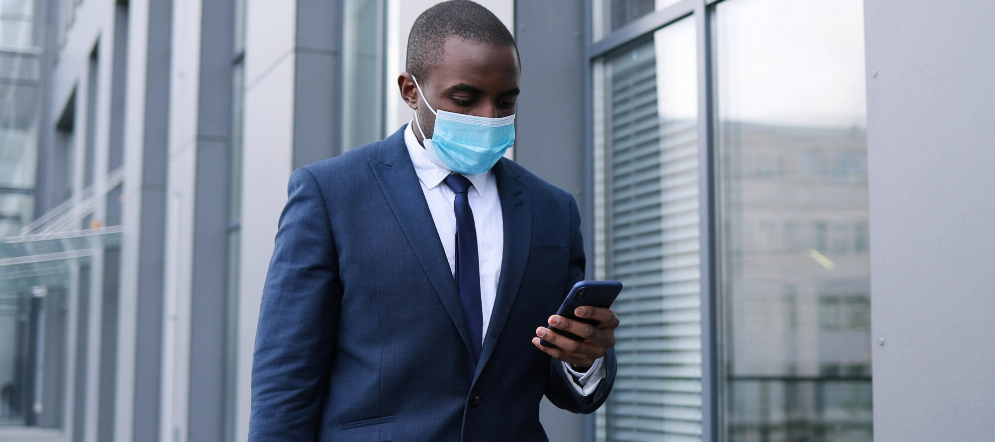 A man in a blue suit and tie, wearing a face mask, checks his smartphone while walking outside a modern glass building, perhaps reviewing visitor management system software on the go.