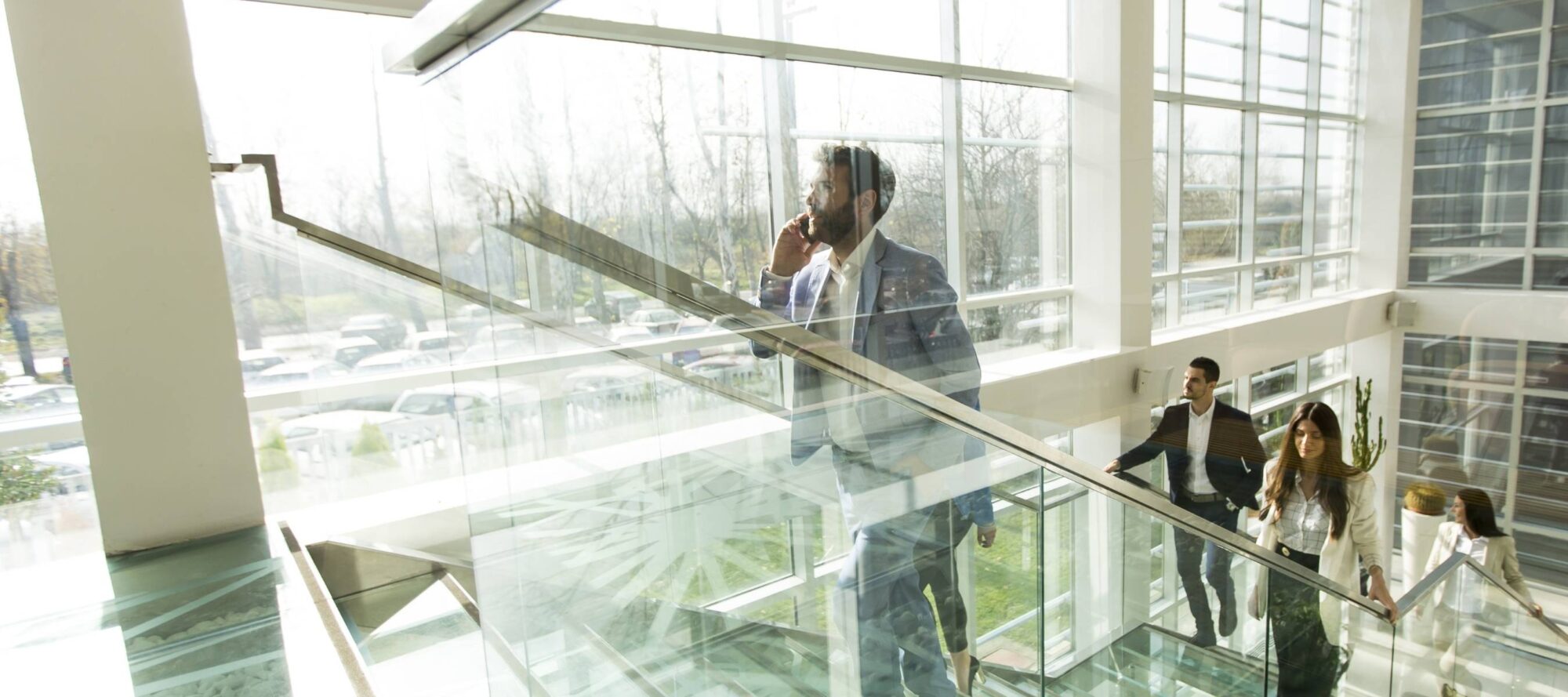 A group of people in business attire walks up a glass staircase inside a modern building with large windows. Sunlight streams in, illuminating the scene. One man in the foreground is talking on a phone, possibly discussing access control—a vital feature of the latest tenant amenity apps.