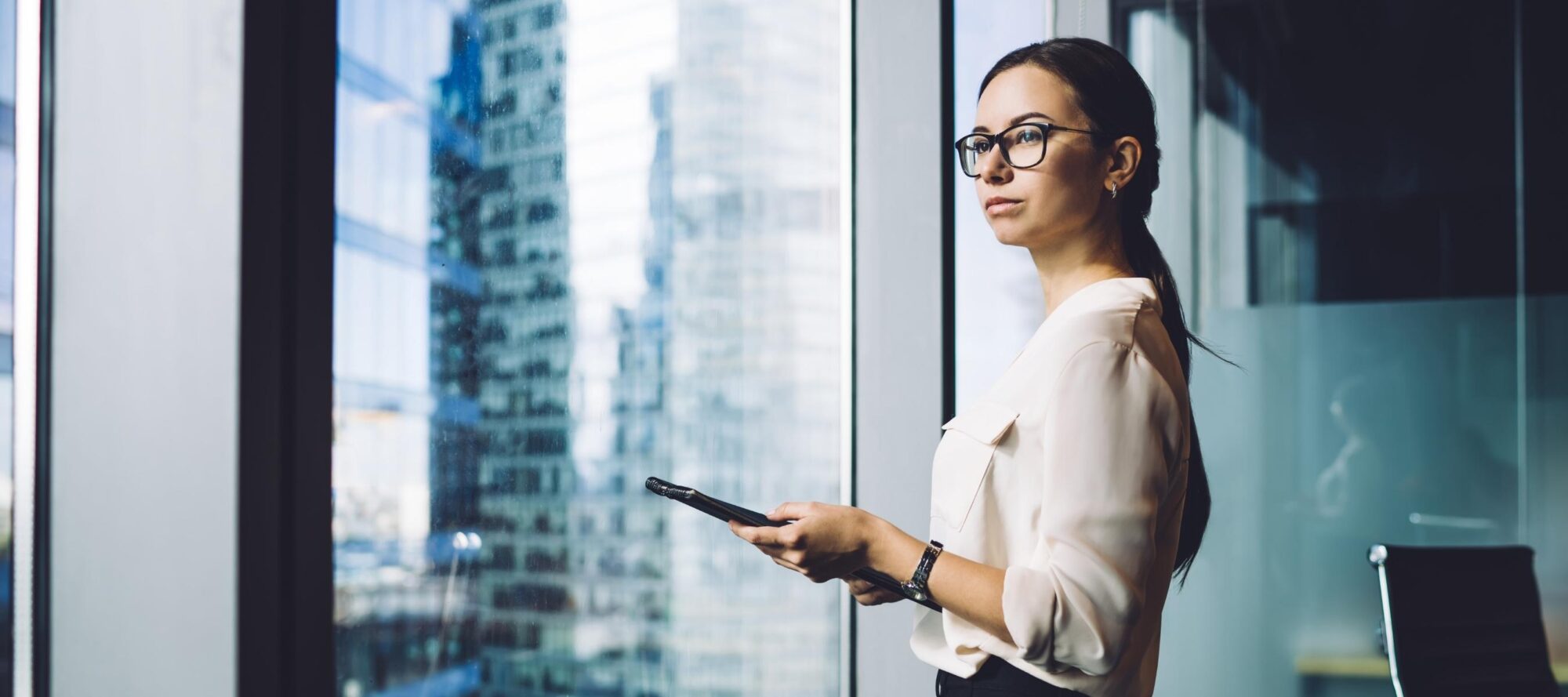 A woman in business attire stands by a large window with a tablet in hand, contemplating what is access control. She wears glasses and has long dark hair, while the cityscape's modern buildings reflect in the glass behind her.