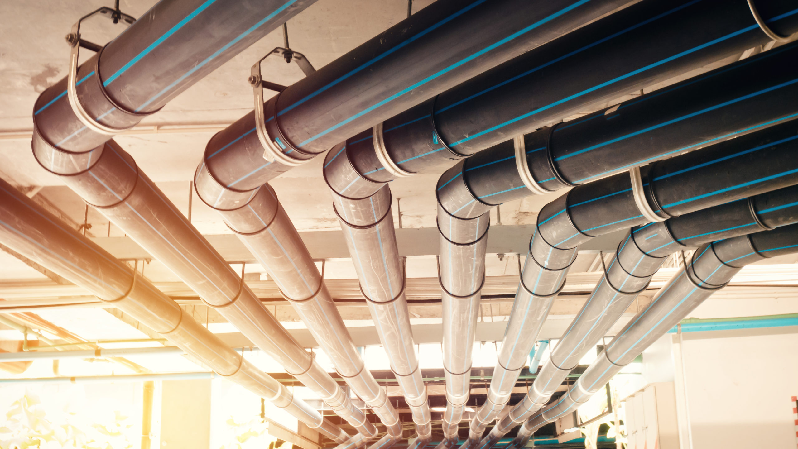 A series of metallic pipes with small blue lines run parallel to each other along the ceiling, part of an efficient HVAC system. The pipes are evenly spaced and suspended by metal brackets, with sunlight filtering in from the left, casting a warm glow that syncs perfectly with its meticulous scheduling.