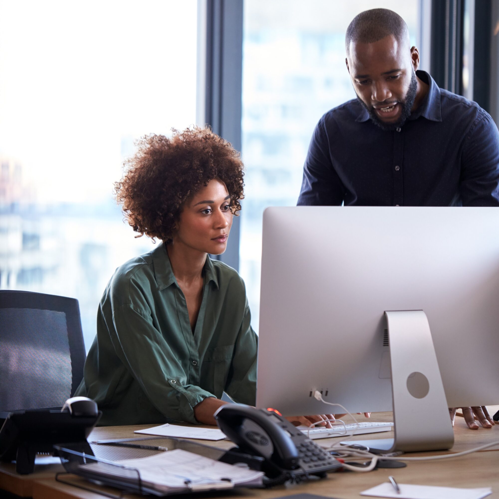 Two colleagues in an office work on a computer. A woman with curly hair, focused on the screen, partners with a man in a dark shirt gesturing towards it. The desk is cluttered with papers, a phone, and a monitor.