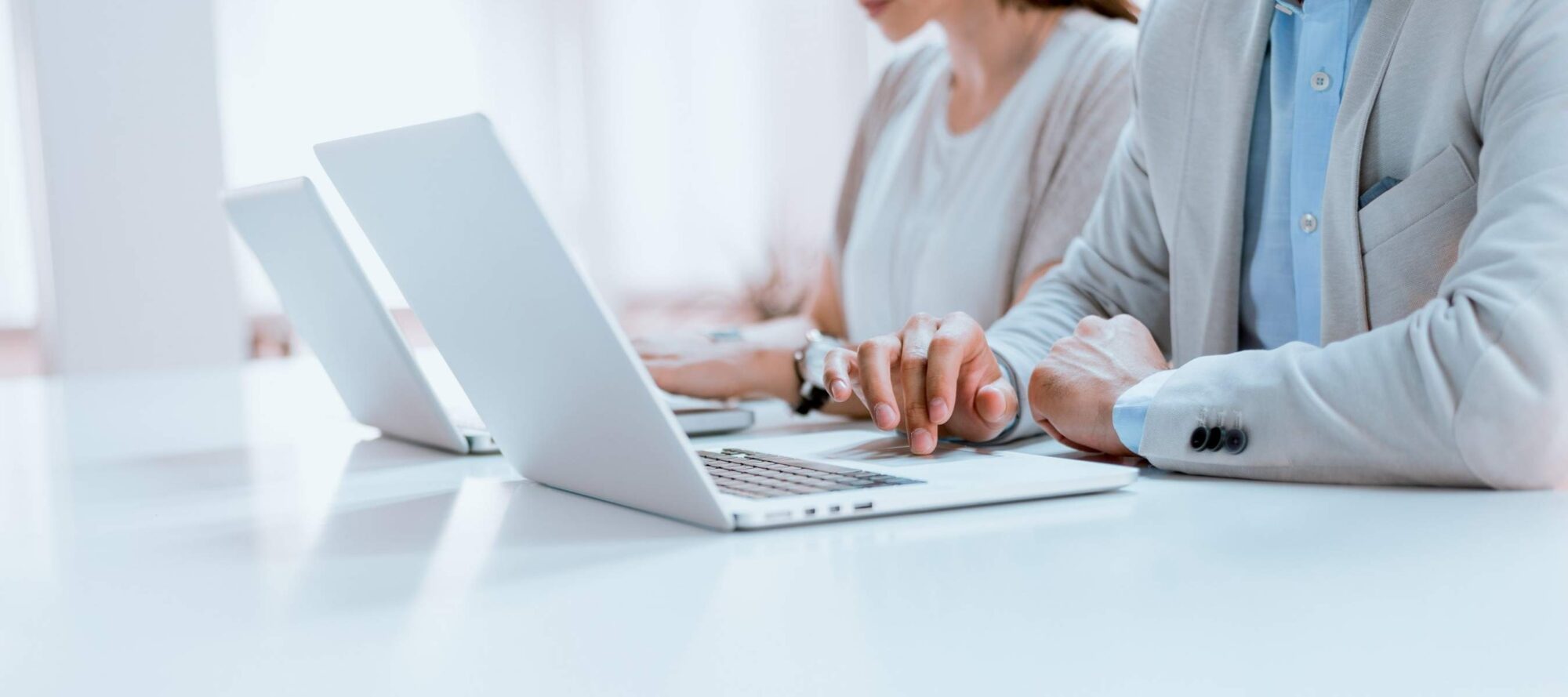 Two people sit side by side, immersed in their laptops, with hands typing efficiently as if fueled by cloud computing accelerated processes. Dressed in business attire, they exude professionalism. The background is bright and softly blurred, highlighting the focus on their work.