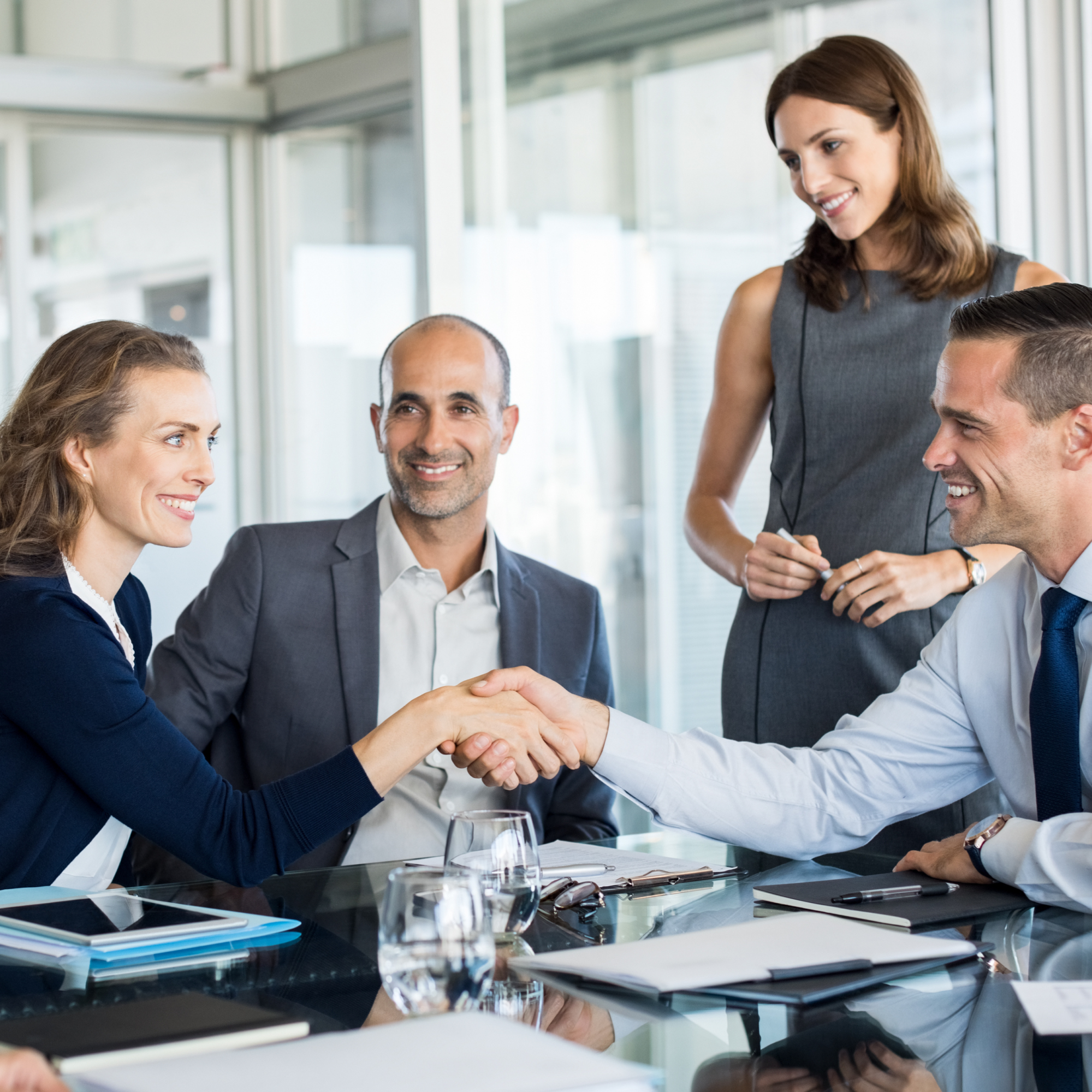 A group of business professionals gathers in a meeting room. A woman and a man, possibly Genea partners, are shaking hands across a glass table, smiling. Three others stand or sit nearby, engaged and watching the handshake. Office supplies and documents are scattered on the table.