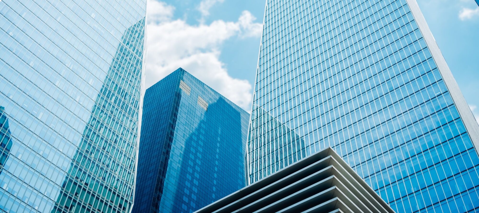 Looking up at a group of modern skyscrapers with glass facades reflecting the blue sky and clouds, one can't miss the changes in access control seamlessly integrated into the design. The architecture features geometric shapes and tall structures, creating a dynamic urban scene.