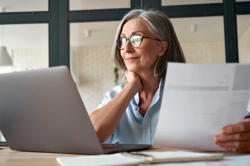 A woman with gray hair and glasses sits at a desk, holding a document in one hand while reviewing tenant billing software on her laptop. She appears thoughtful and content. The room has large windows and a modern design.