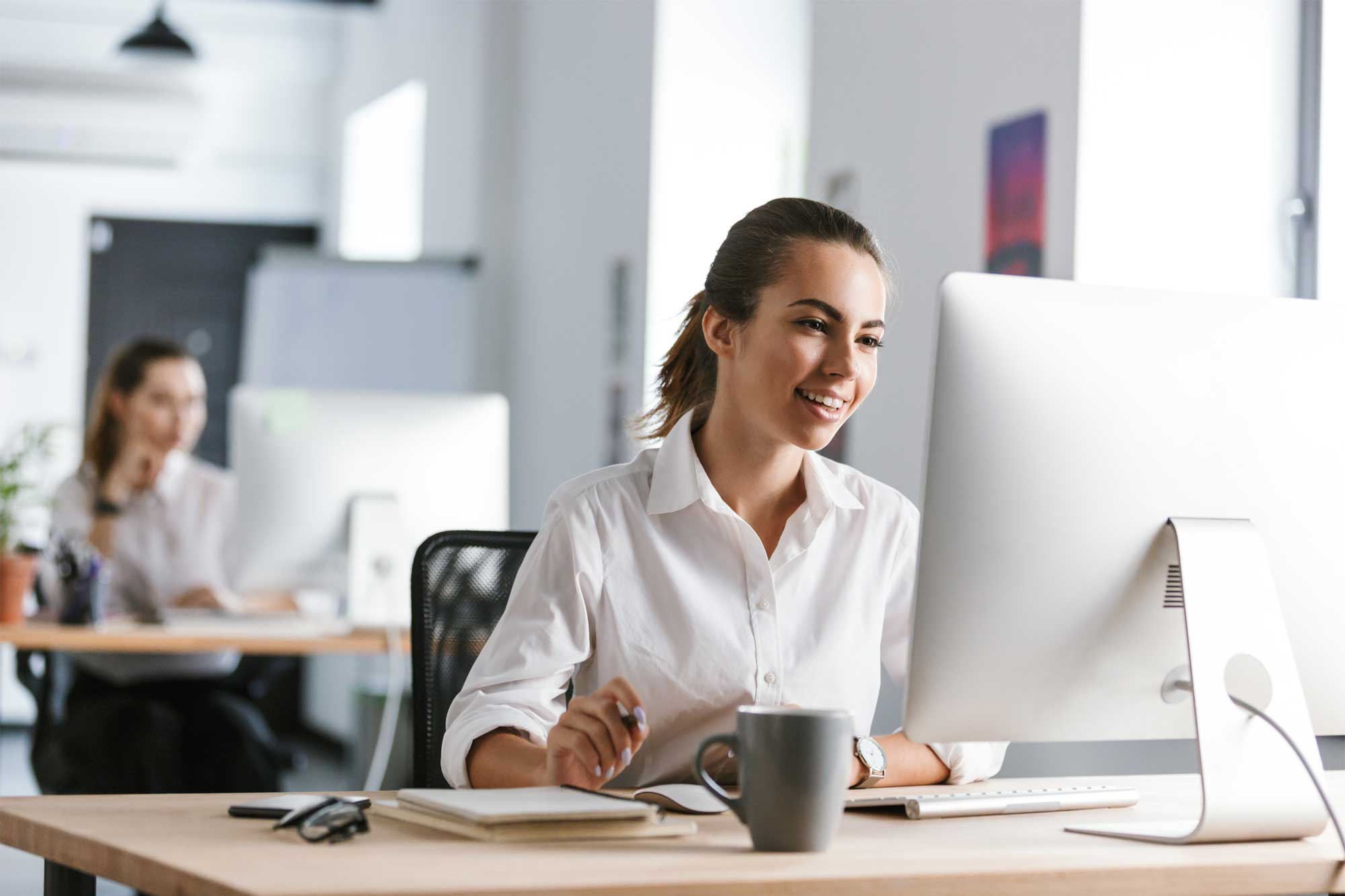 A woman in a white shirt is smiling as she works on tenant billing software at her desk. A coffee mug and notebook are nearby. In the background, another person is sitting at a desk in a modern, well-lit office space.