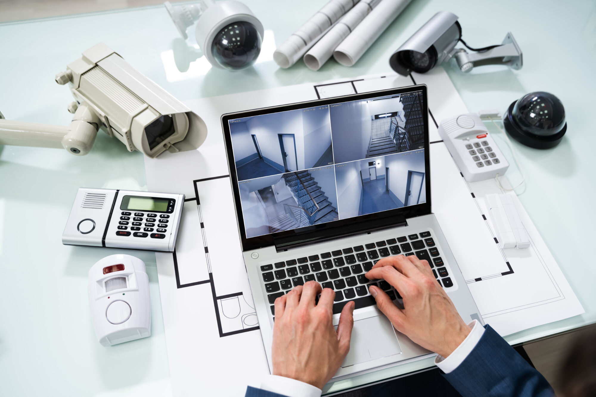 A person in a suit types on a laptop displaying CCTV feeds, expertly navigating video management software. The desk is cluttered with security cameras, blueprints, and electronic devices, suggesting a focus on surveillance and security systems.