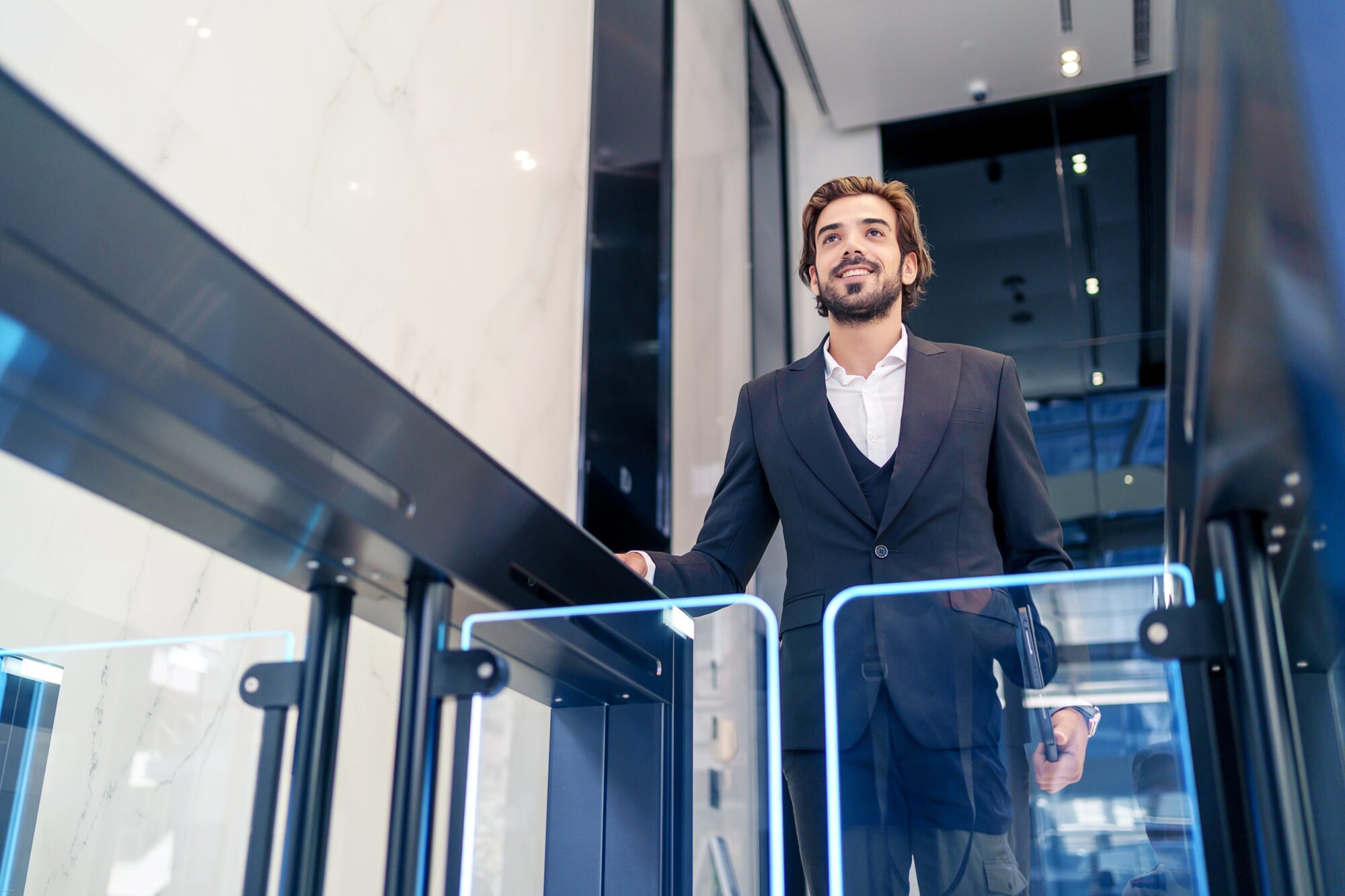 A man in a business suit smiles as he walks through the state-of-the-art entrance gate, seamlessly integrated with advanced physical security features. The office building boasts sleek polished surfaces and bright lighting, reflecting its modern and secure design.