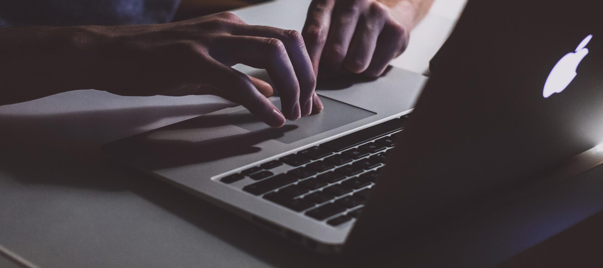 In a dimly lit setting, a person types on a MacBook with softly glowing keys. The focus is on their hands and the laptop, creating a moody ambiance as FIDO2 Authentication ensures seamless security in the background.