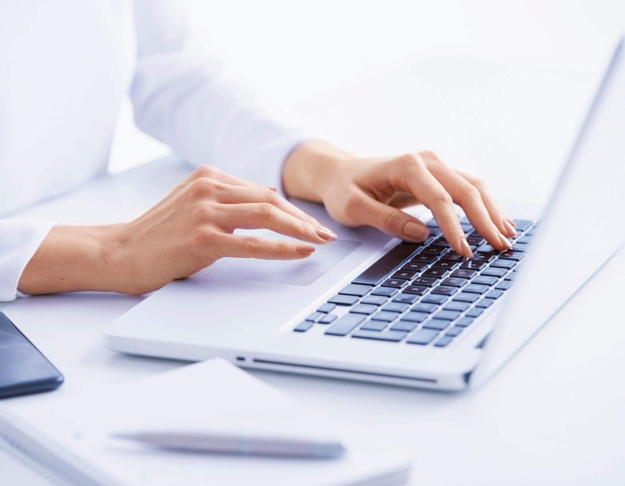 Close-up of a persons hands typing on a laptop keyboard. The setup includes a smartphone, a notebook, and a pen on a white desk. The background is softly blurred, and the scene conveys a sense of focus and productivity.