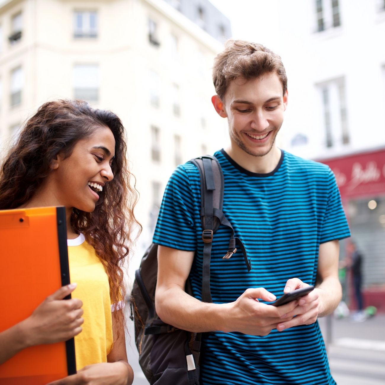 A man and woman are happily walking on a city street, looking at a smartphone. The woman, in a yellow shirt, holds orange folders while the man, wearing a striped blue shirt and carrying a backpack, discusses access control for schools with her.