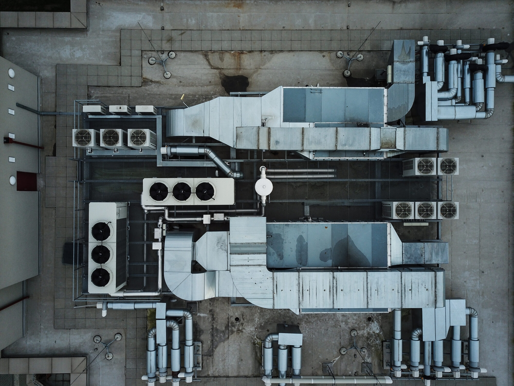 Aerial view of a building's rooftop reveals a data and analytics-driven layout of complex ventilation and air conditioning systems. Multiple ducts and units form an intricate pattern of metal pipes and rectangular structures, strategically arranged on the concrete surface.