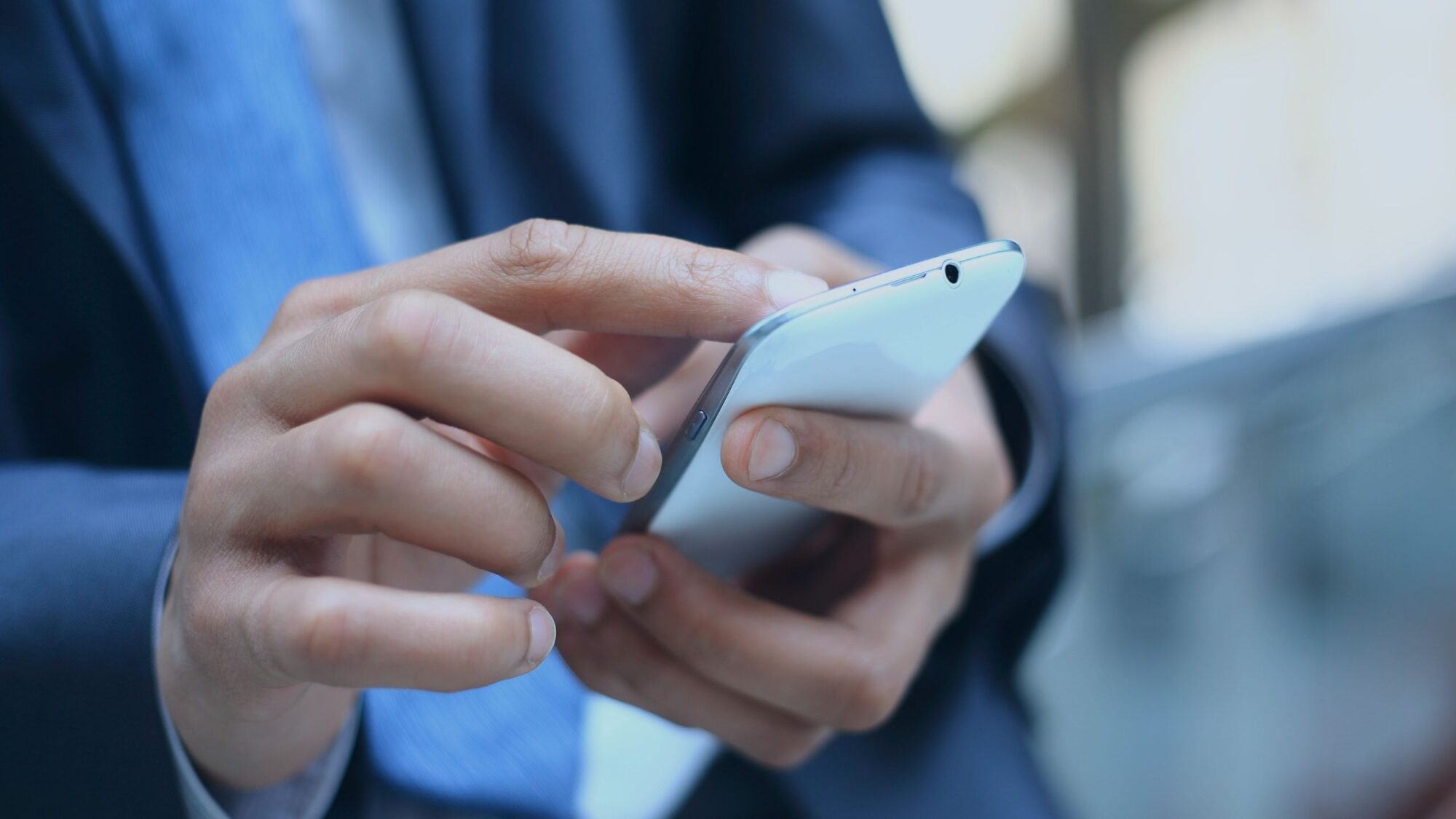 A person in a suit is using a smartphone. Their hands are focused on typing or scrolling on the device. The background is blurred, highlighting the action and the device.