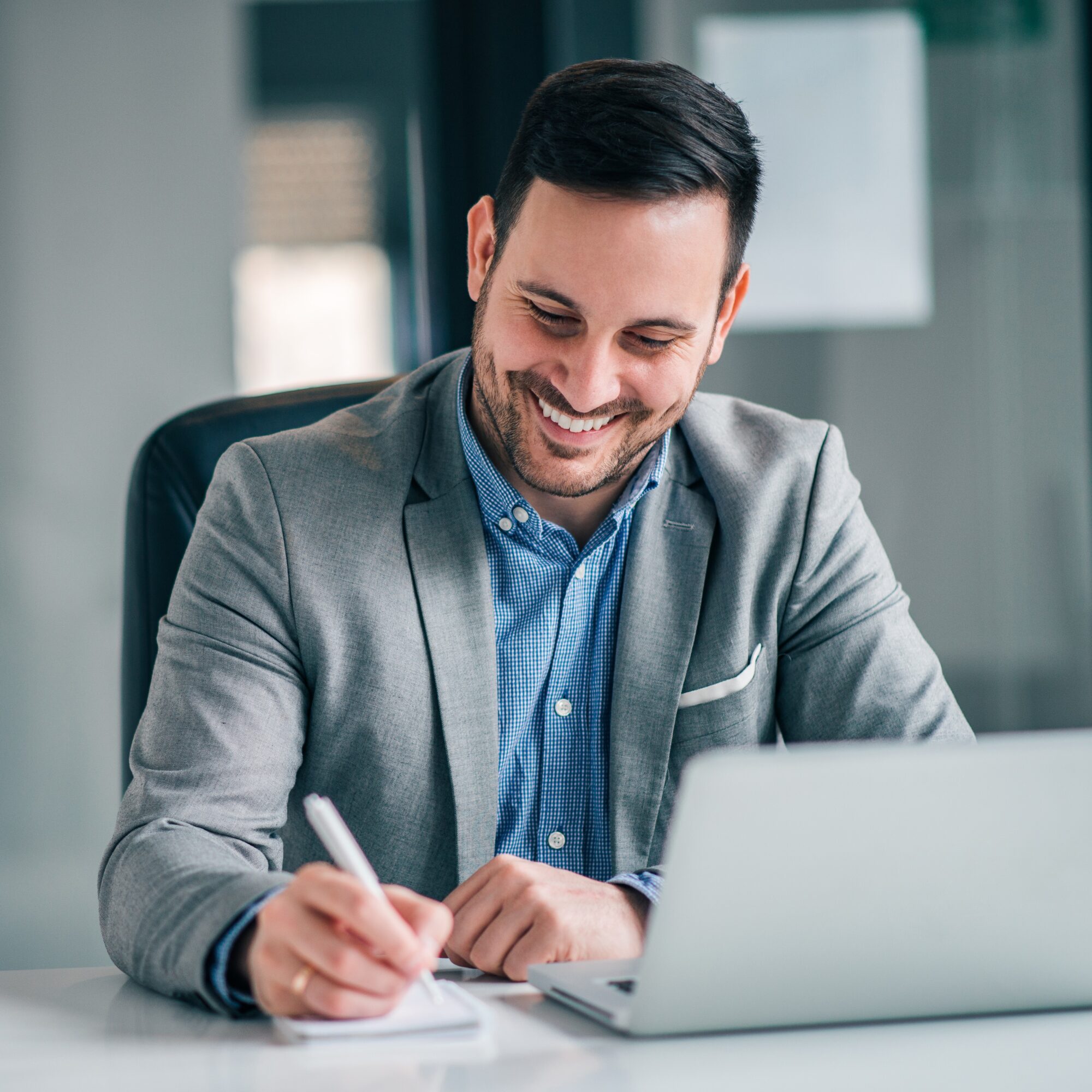 A man in a gray suit and blue shirt is seated at a desk, smiling as he writes on a notepad. With a laptop in front of him, he appears to be in an office setting, possibly strategizing plans for advanced commercial security systems.