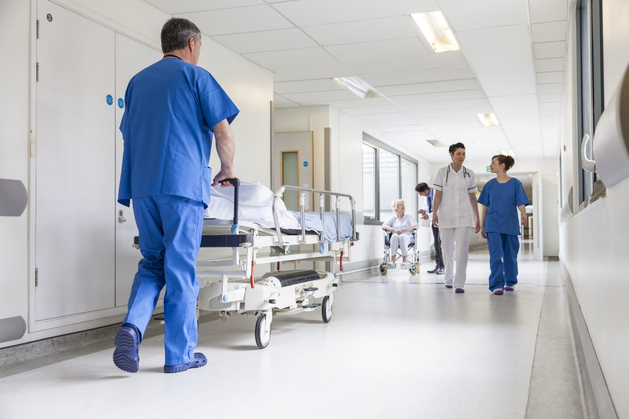 In the bright, clinical hospital hallway, a person in blue scrubs pushes a hospital bed past access-controlled doors. Two people in white coats walk and talk, while another sits in a wheelchair, all demonstrating the seamless flow of healthcare professionals within this secure environment.