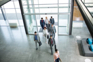 A group of nine professionally dressed people walk through the lobby of a modern office building. The space features large windows, natural light, and sleek furniture, seamlessly integrating a visitor checkout area that maintains the elegant aesthetic.