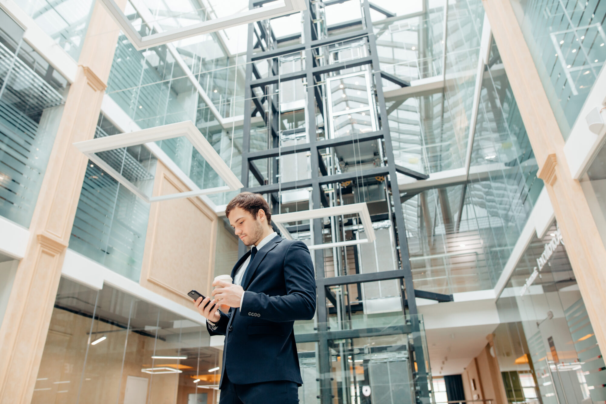 A man in a suit stands in a modern, glass-walled atrium, holding a smartphone and a coffee cup. Above him is an exposed elevator shaft equipped with sleek access control technology. The space is filled with natural light and showcases impressive architectural design.