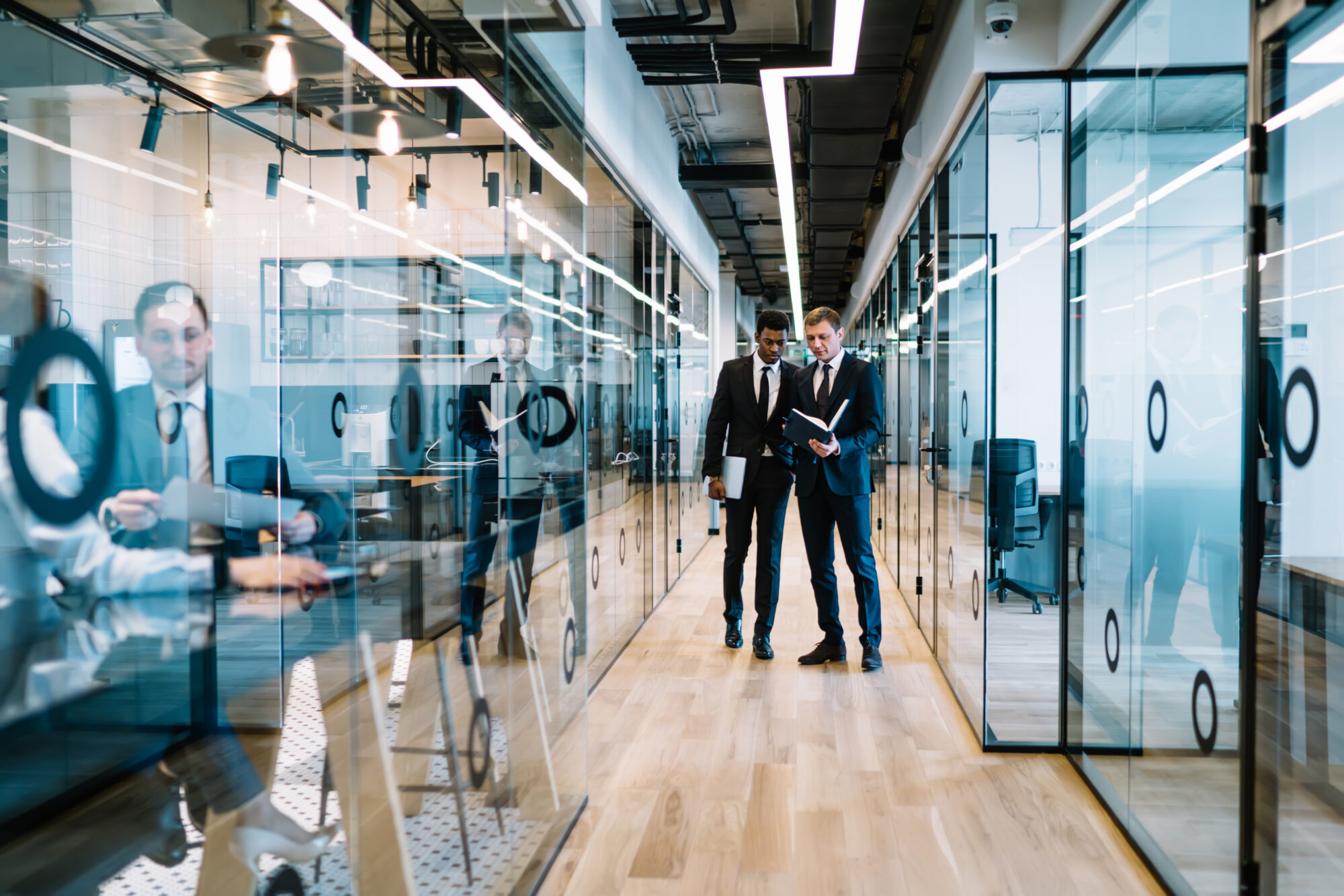 Two men in suits walk through a modern smart office hallway with glass walls. One reads a document while holding a tablet, and the other listens attentively. Reflections of colleagues working at desks are visible through the glass.