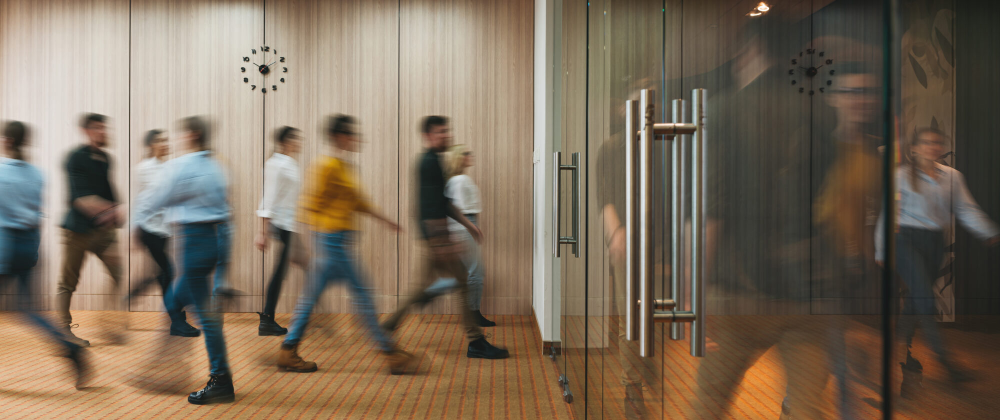 A group of people walking briskly in a modern office corridor with non-proprietary access control, a glass door in the foreground, and a wooden wall with a clock in the background. The image captures motion blur, indicating a busy atmosphere.