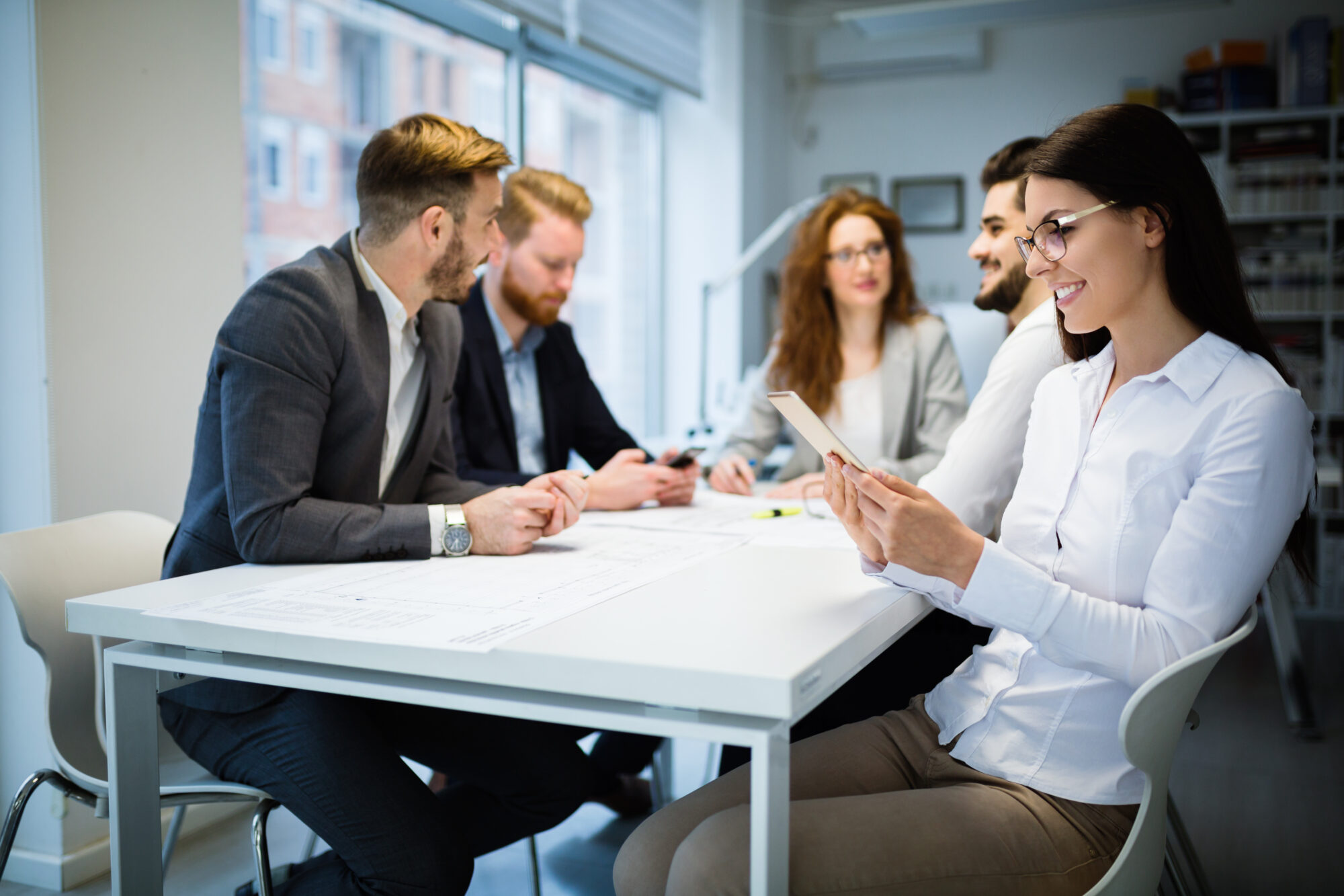 A group of five people sits around a white table in an office setting. Four focus on documents, while one woman, smiling and wearing glasses, explores Vertx insights on a tablet. Large windows in the background provide natural light.