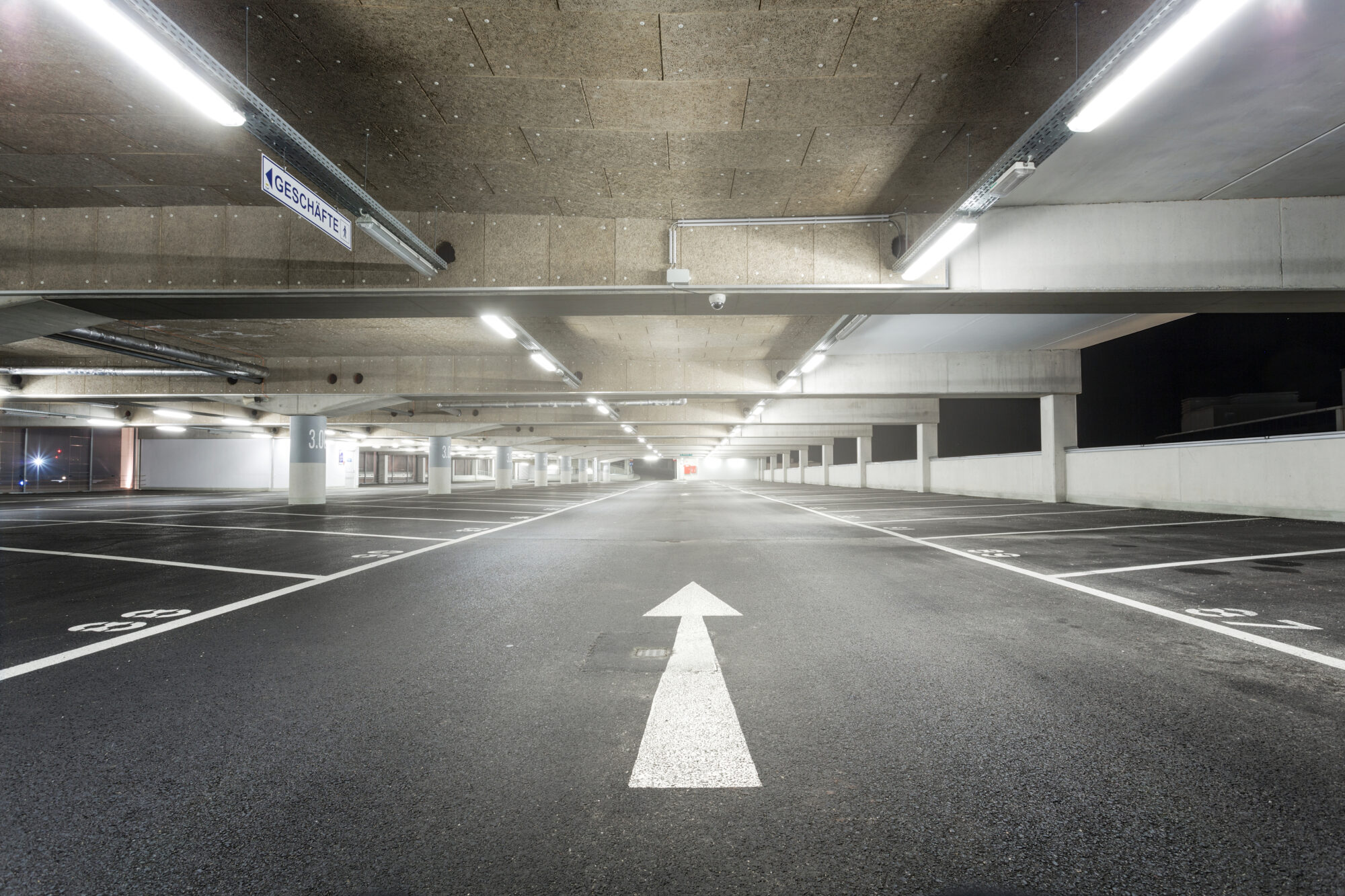 A well-lit, empty multi-level parking garage features marked spaces and a forward-pointing arrow on the asphalt floor. Fluorescent ceiling lights illuminate the area while a sign reading Kassenautomat is visible on the left, embodying the efficiency of flash parking.