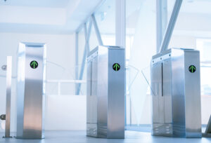 A row of three sleek, metallic turnstiles with glass panels stands in an open, modern building interior, serving as a sophisticated physical access control system. Green arrow indicators on each turnstile suggest they are ready for entry. Sunlight filters through large windows, creating a bright atmosphere.