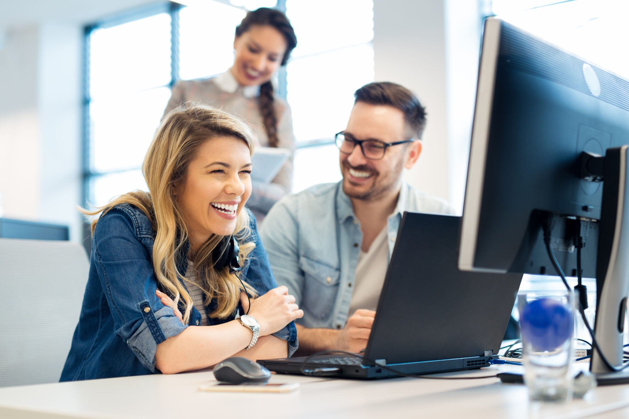 Three people in a bright office smile while looking at a computer screen. Two are seated at a desk with laptops, ensuring access control, while the third person stands behind them. The atmosphere is cheerful and collaborative, as ideas flow freely in this dynamic workspace.