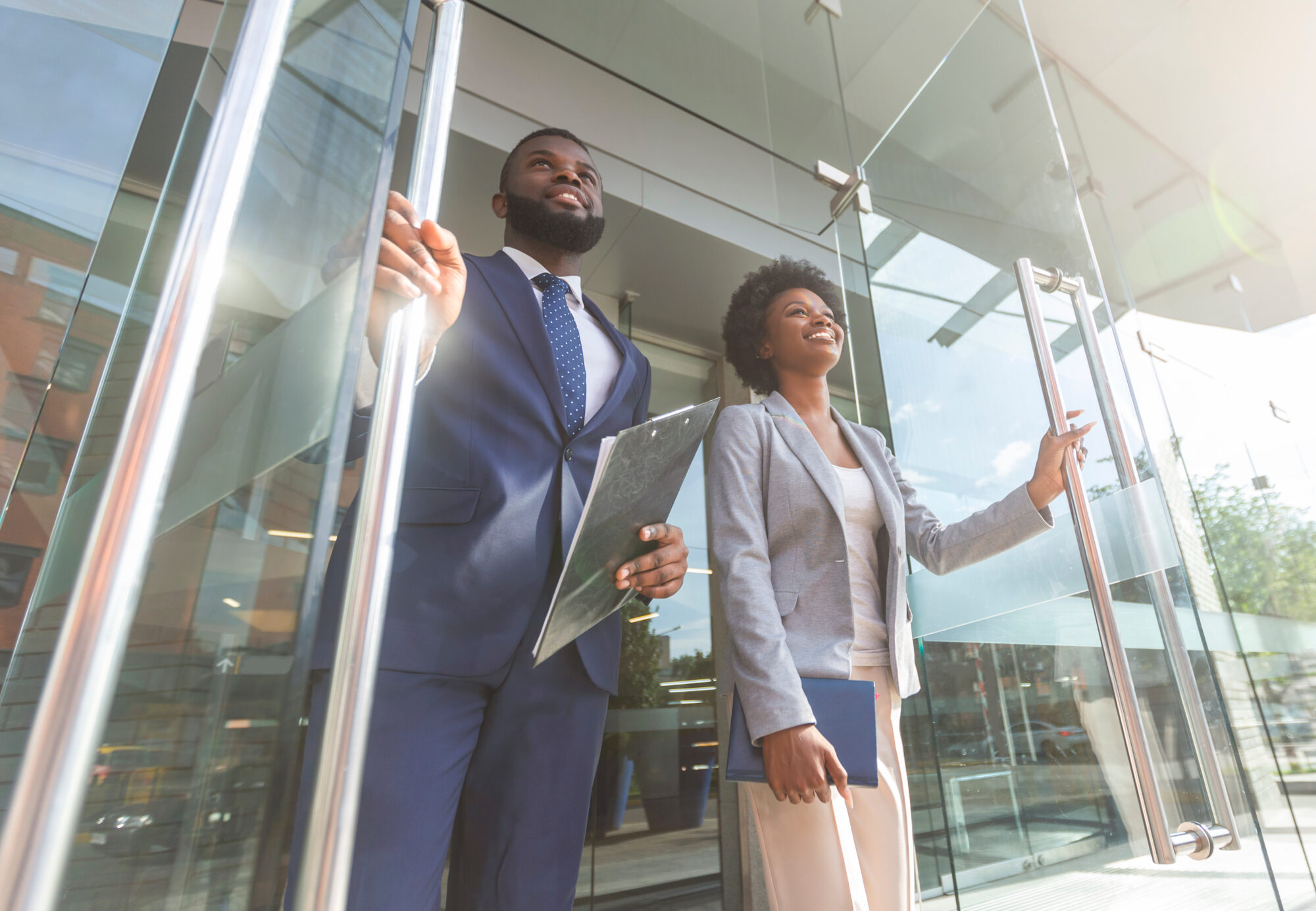 A man and woman in business attire are exiting a modern glass building. The man holds a clipboard, possibly containing physical security checklists, while the woman carries a folder. Both are smiling and looking ahead, suggesting a professional or successful moment.