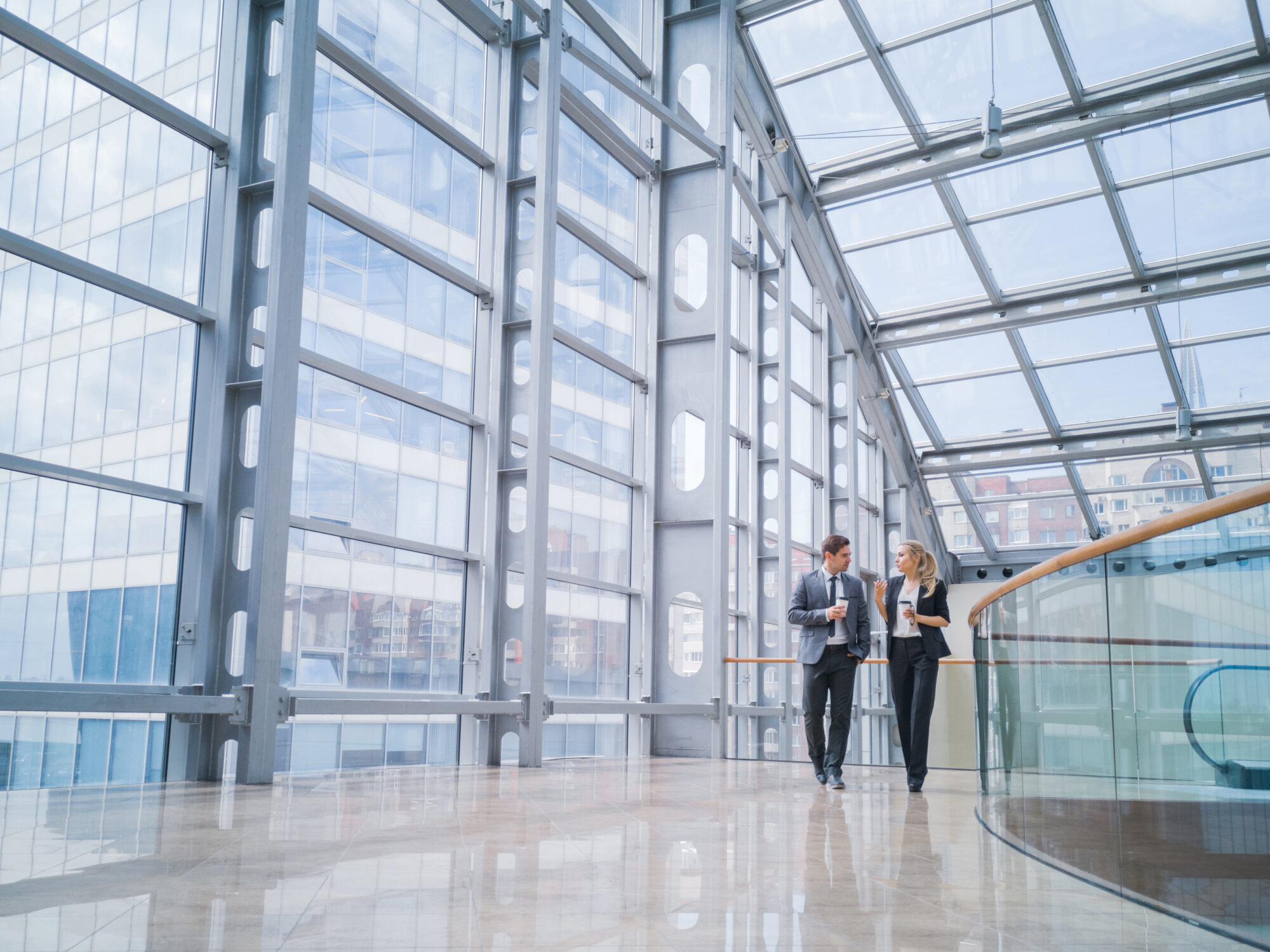 A man and a woman are walking and talking in a spacious, modern building with large glass windows and a high ceiling. They both hold paper cups, engrossed in conversation about wireless access control. The shiny floor reflects the natural light streaming in.