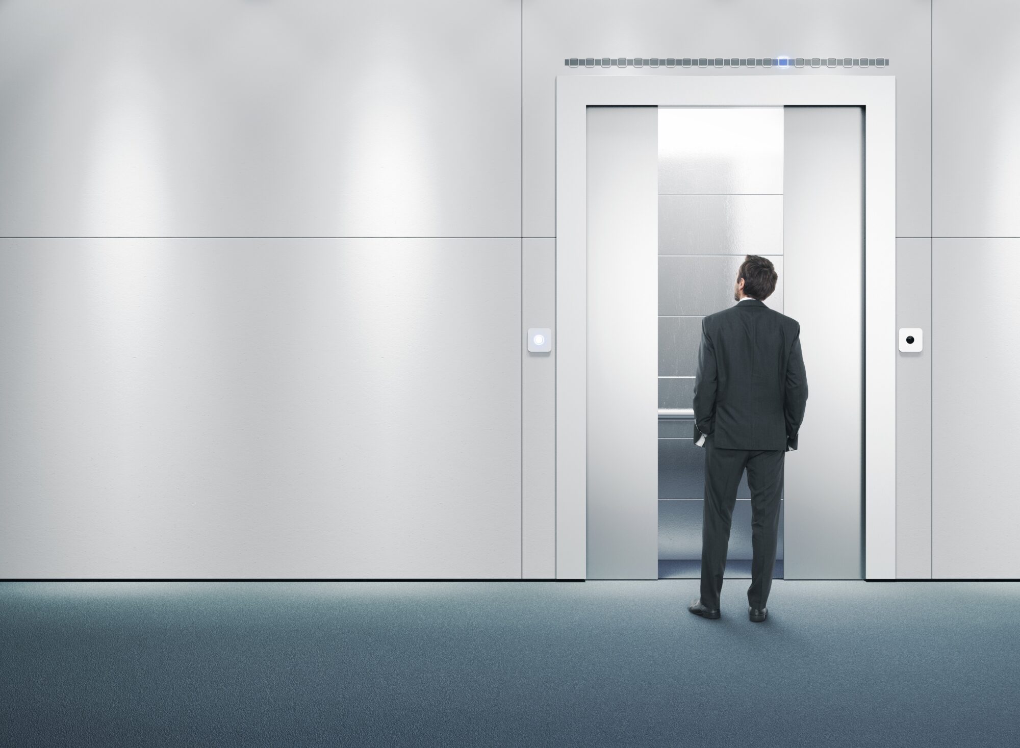 A man in a suit stands in front of an open elevator with a modern, minimalist design. Enhanced by elevator access control, the atmosphere is secure yet inviting. The grey walls and blue carpet floor are well-lit, creating a professional setting.