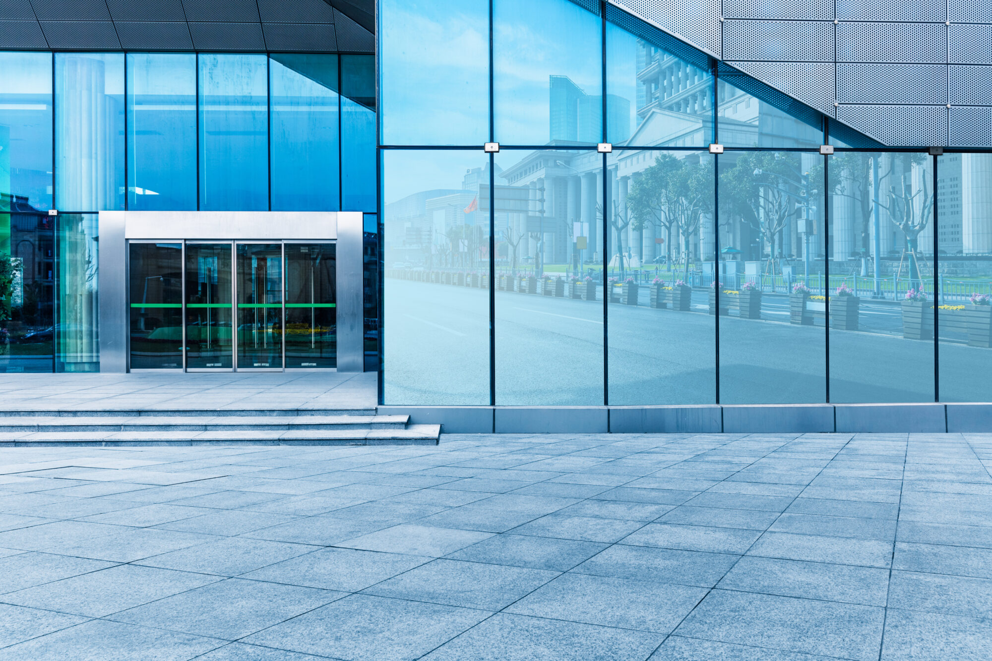 The modern building exterior, outfitted by security integrators, boasts large glass windows reflecting an urban landscape. Its entrance features sliding glass doors, while the foreground reveals a wide, empty stone pavement. The scene is calm and clear on a bright day.