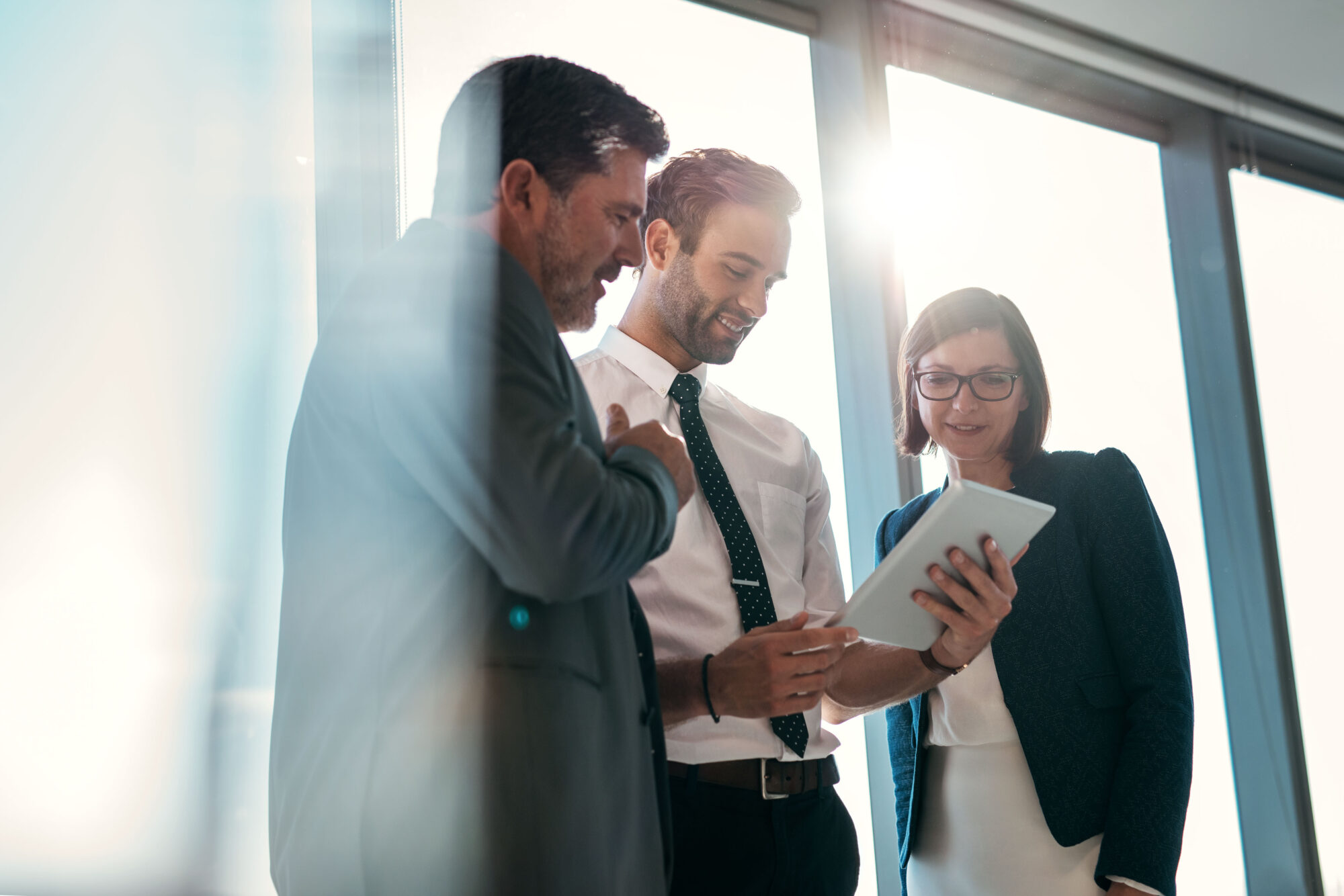 Three business professionals are gathered in a brightly lit office, reviewing muster reports on a tablet. Two men, one wearing a tie, and a woman in glasses actively discuss the details, as sunlight streams through the windows.