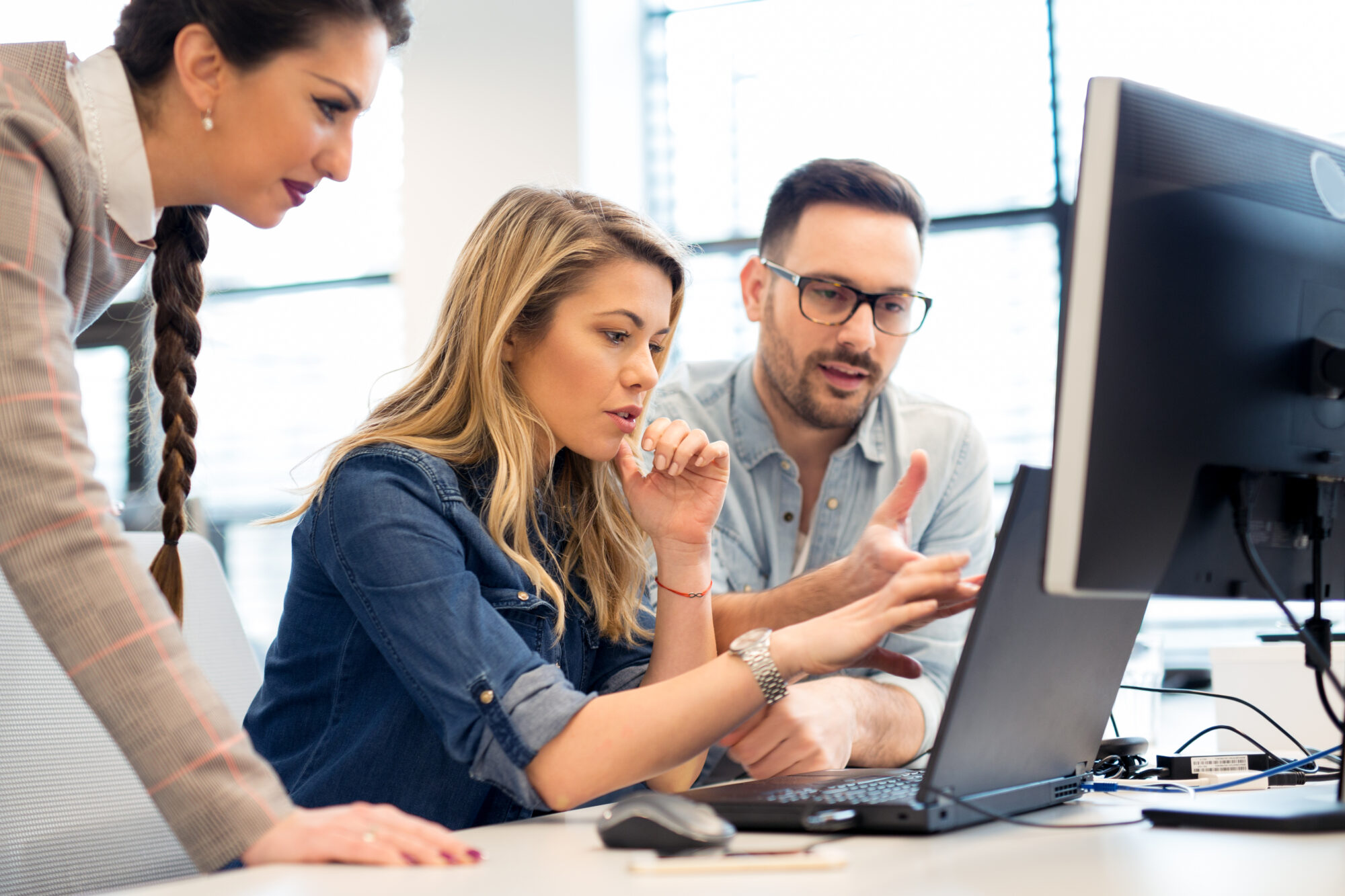 Three people collaborate in an office setting, focused on a computer screen. One person points at the screen while discussing custom attributes with the others. They appear engaged and concentrated on the task at hand.