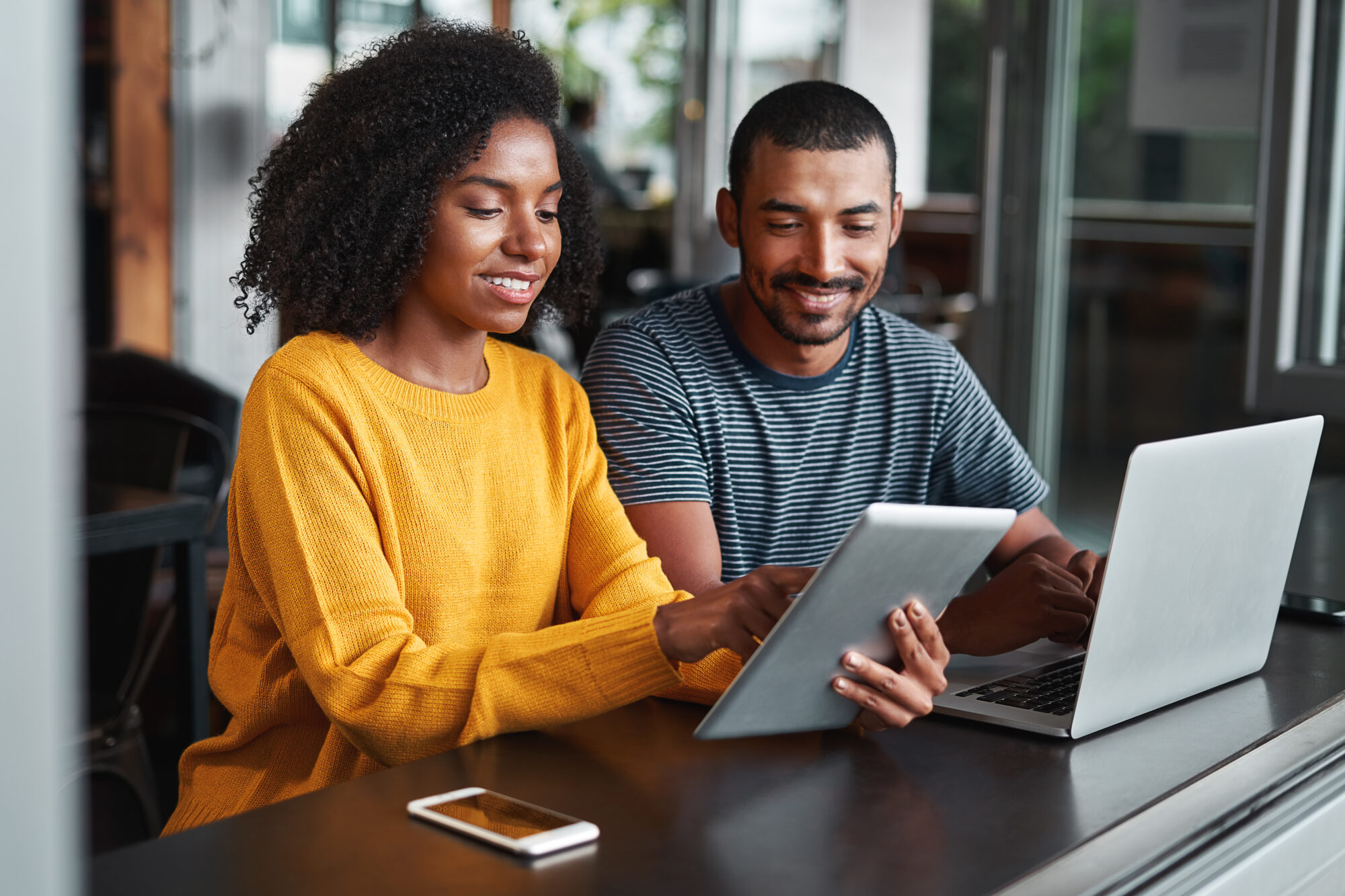 A woman in a yellow sweater and a man in a striped shirt sit at a table, engaged and smiling, as they explore critical event management solutions on a tablet. A laptop is open before the man, while a smartphone lies nearby, underscoring their tech-savvy collaboration.