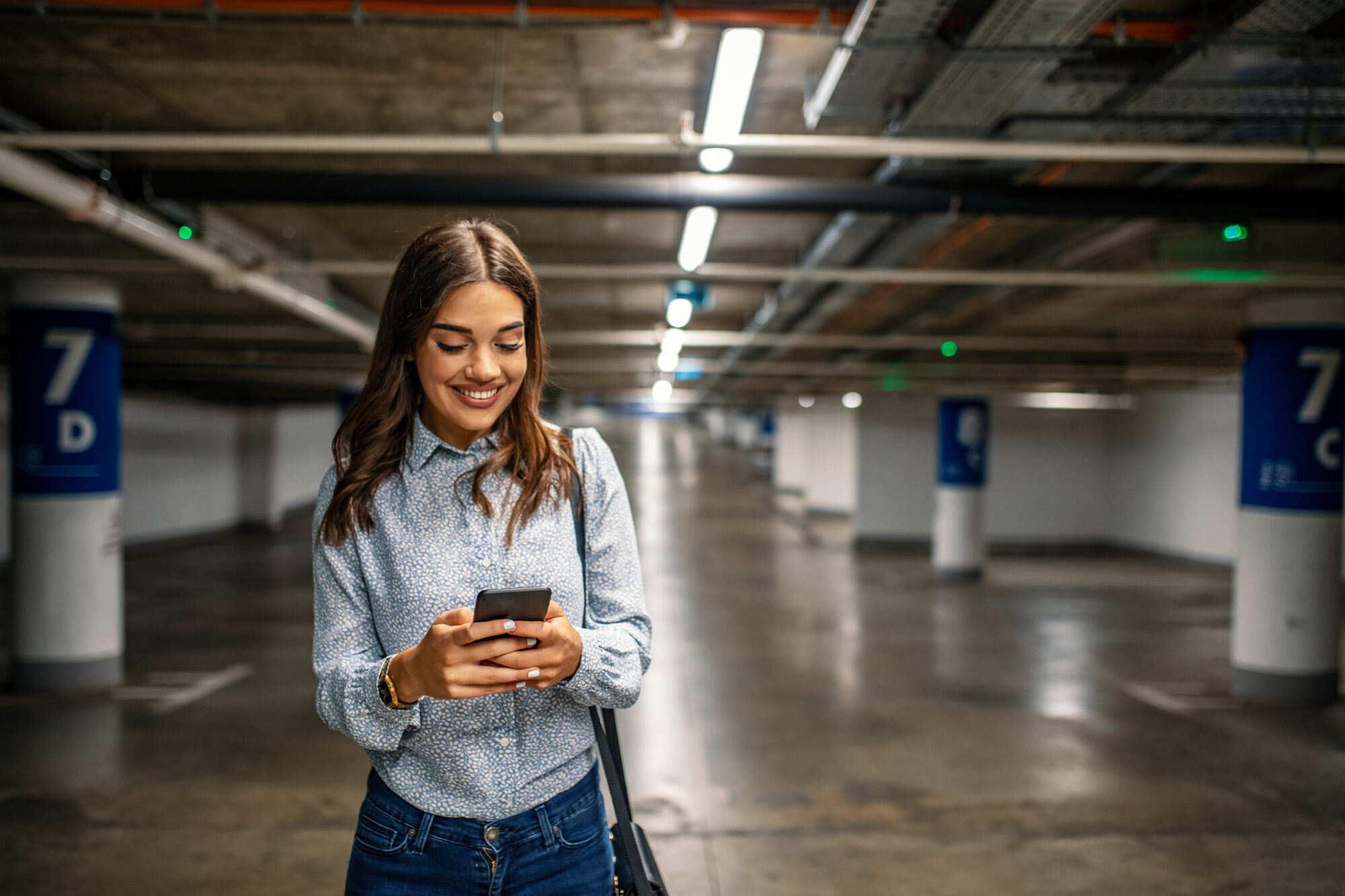 A woman smiles at her phone in an underground parking lot, standing between concrete pillars with numbered signs. Wearing a patterned blouse and jeans, handbag over her shoulder, she seems content, perhaps having just discovered the convenience of parking management software under the bright fluorescent lights.