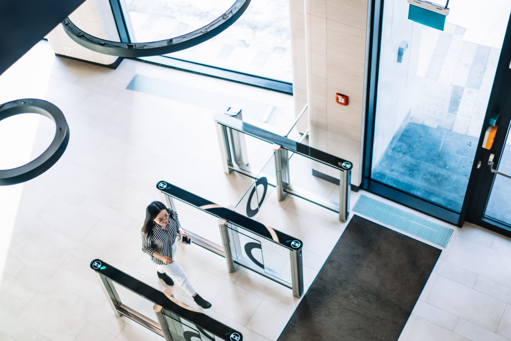 A person walks through a modern security gate installed by Convergint in a bright, tiled office entrance. The area features glass barriers, large windows, and a dark floor mat near the door. The overall environment appears clean and contemporary.