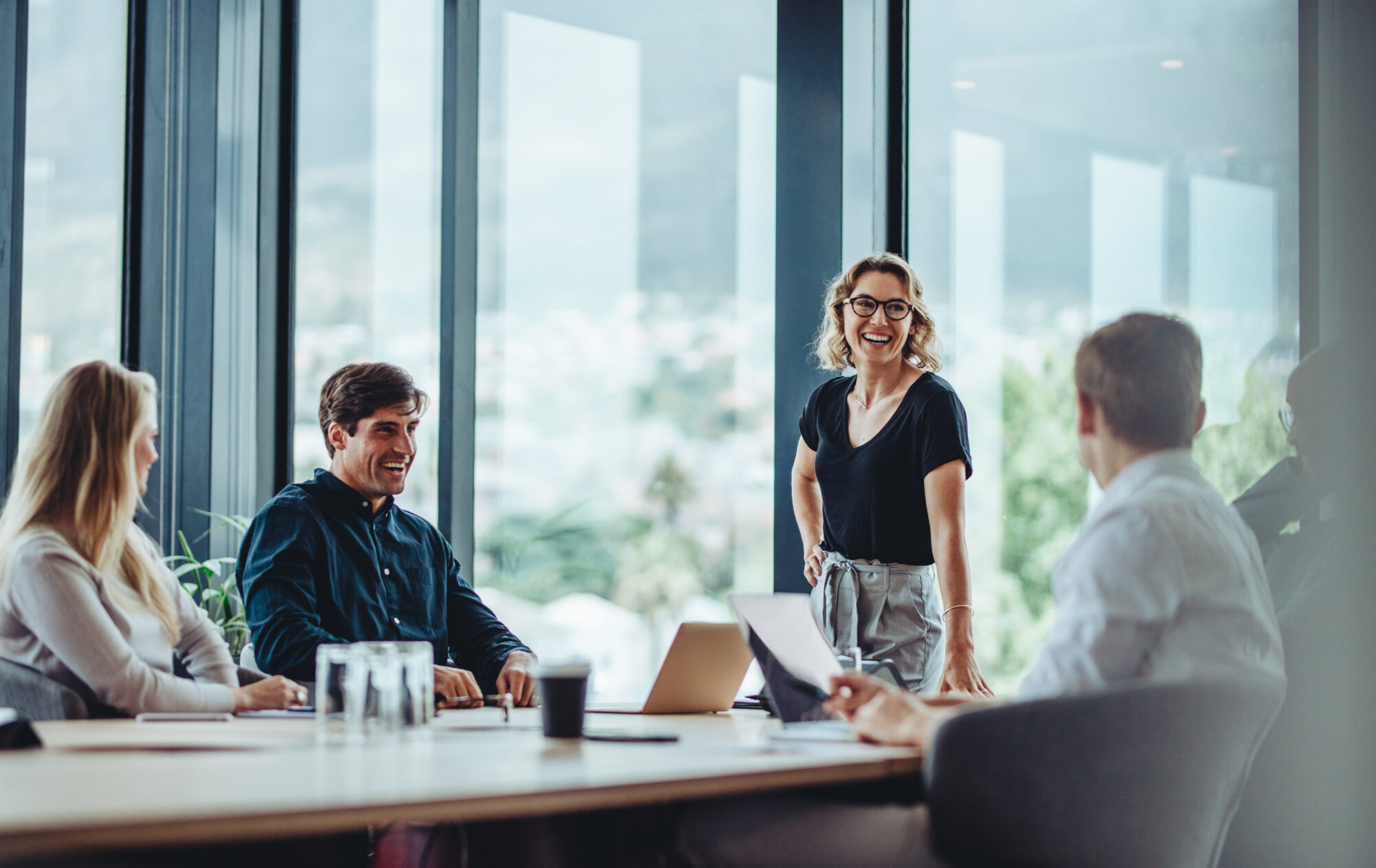 A group of five people in a modern office, smiling and engaged in a meeting. Large windows provide natural light, highlighting the collaborative atmosphere. A woman stands at the head of the table while others sit, suggesting an informal discussion about custom roles within their team.