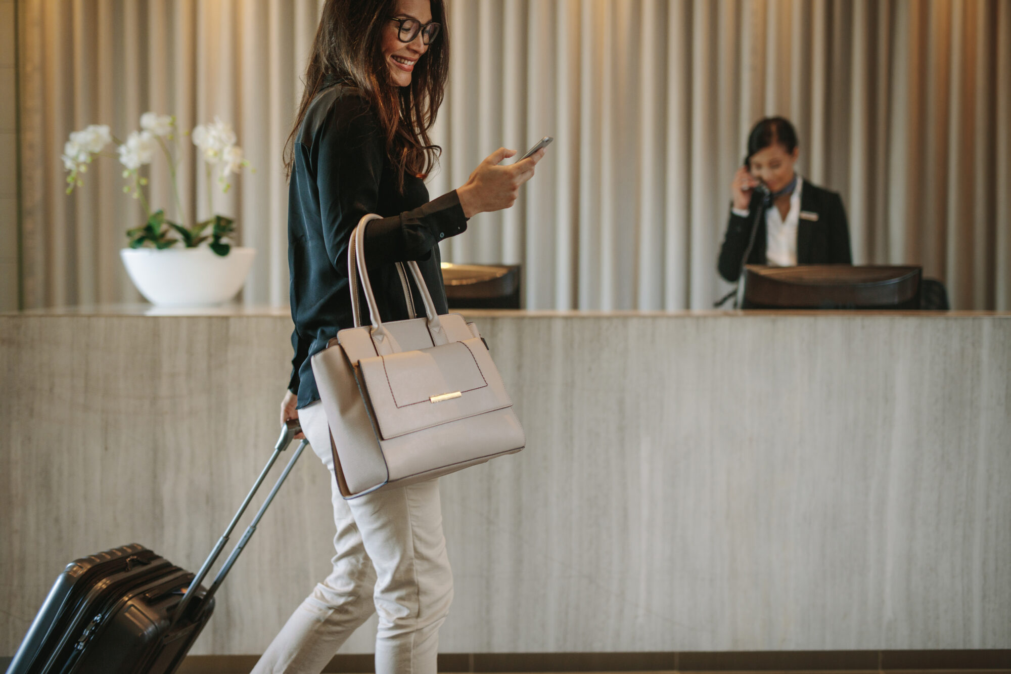 A woman with glasses, pulling a suitcase and holding a smartphone, walks through a hotel lobby. She's dressed in a black shirt and white pants. In the background, the receptionist manages calls while monitoring the hotel's sophisticated access control system.