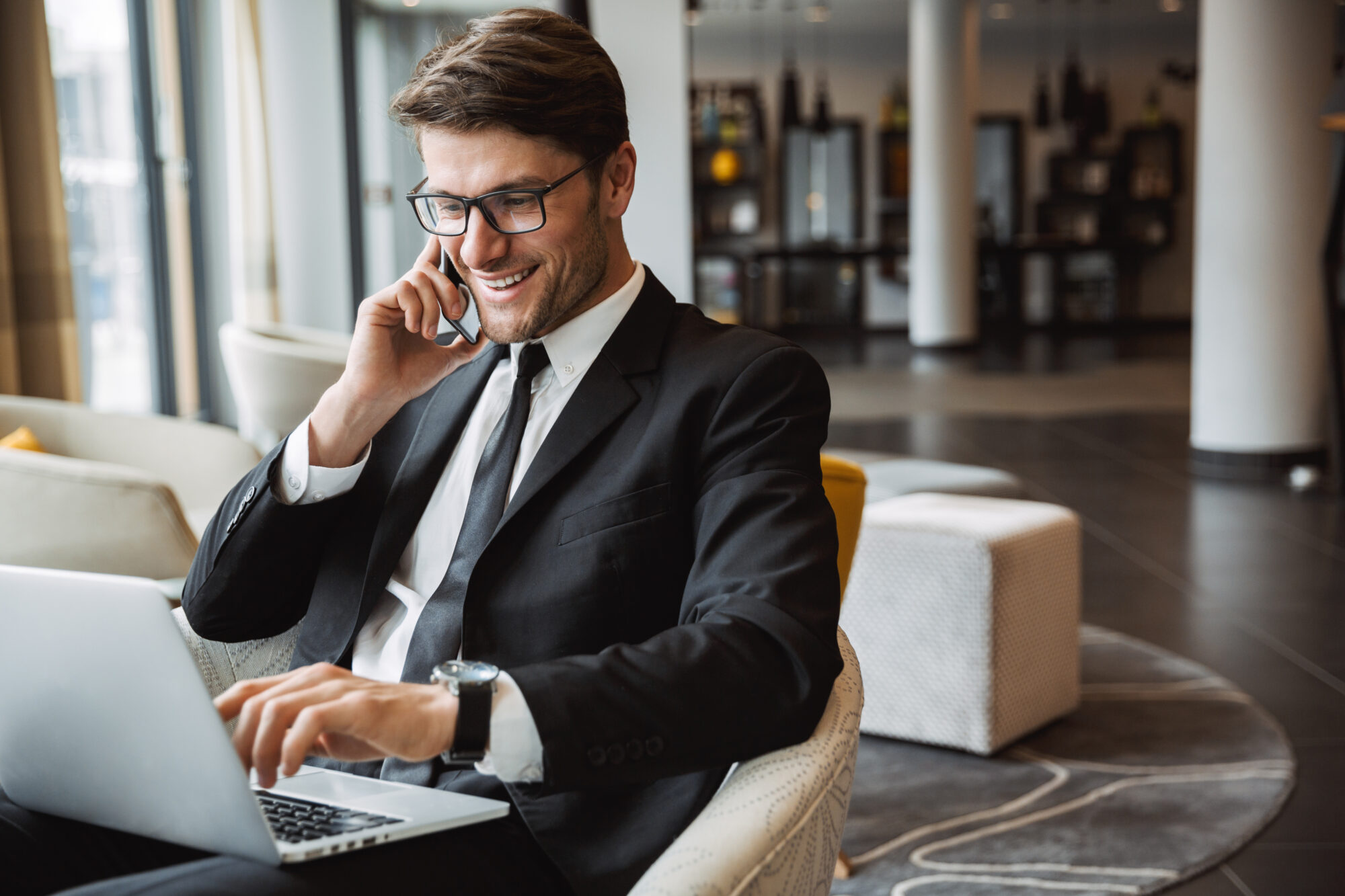 A man in a black suit and glasses sits in a modern office, smiling while talking on a phone. He's working on a laptop, managing settings for the hotel access control system, with contemporary decor visible in the background.