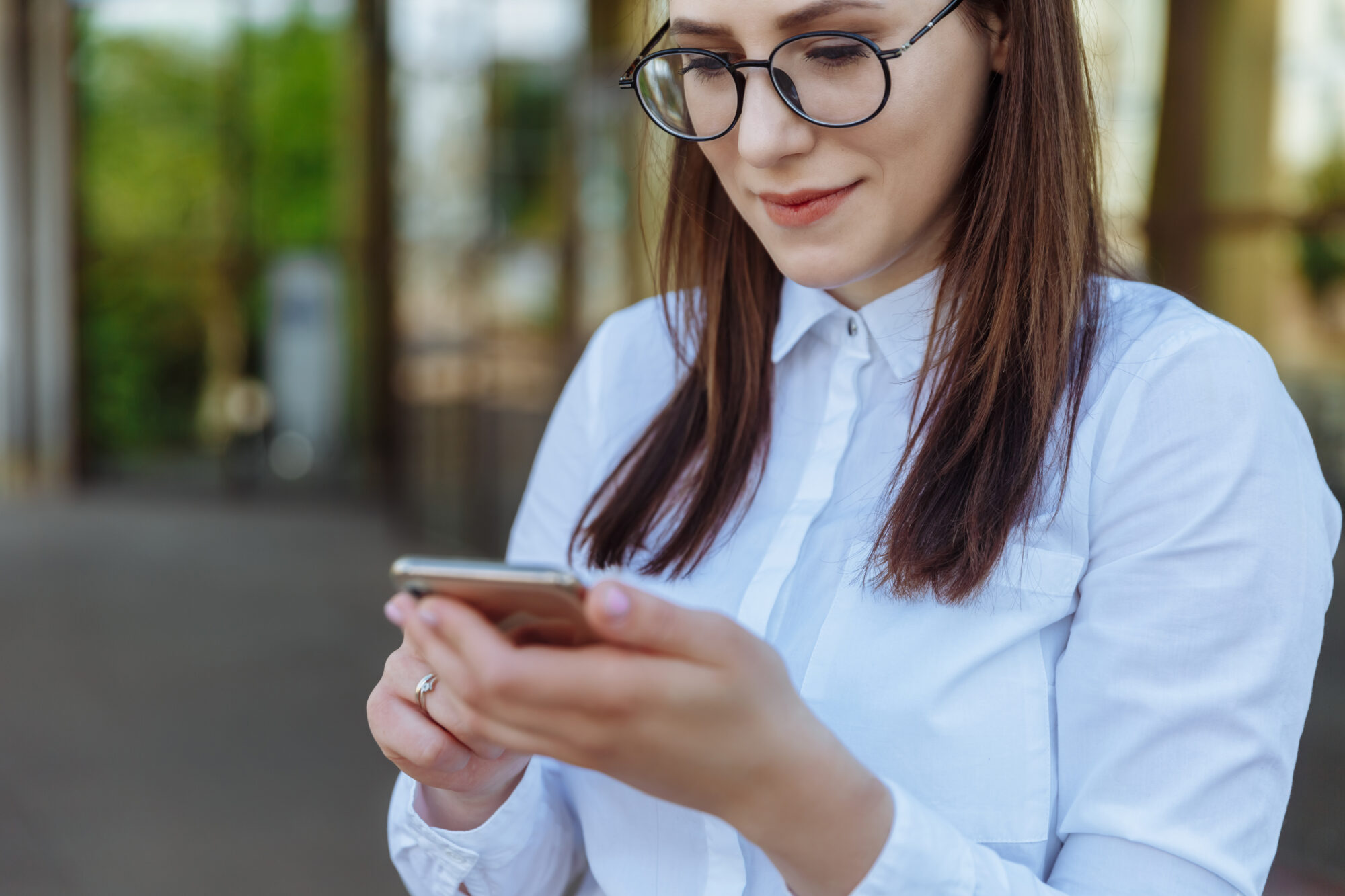 A person with long brown hair and glasses is wearing a white shirt and holding a smartphone, immersed in managing their Apple Wallet. They appear focused on the screen, while the blurred background hints at an outdoor setting.