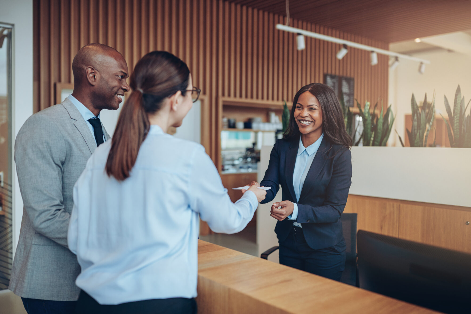 Three business professionals are interacting in an office setting. A woman in a suit is smiling and shaking hands with another woman across a reception desk, discussing the new hotel access control system, while a man stands beside them, also smiling. The background features wooden decor and plants.