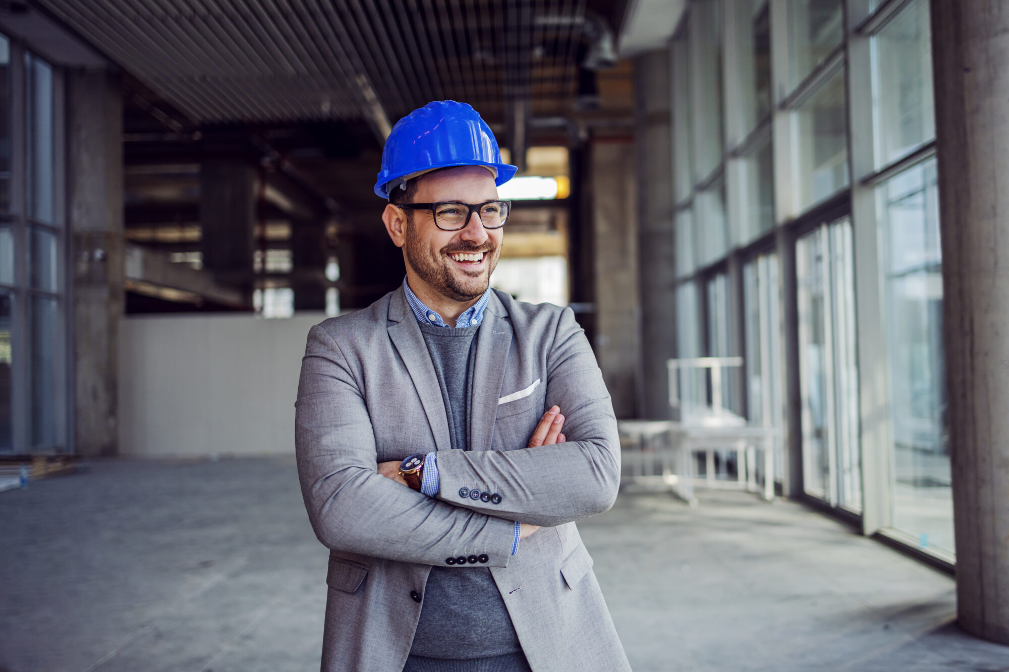 A smiling man in a gray suit and blue hard hat, embodying the expertise of building engineers, stands with arms crossed inside a modern building under construction. Large windows and unfinished interior walls are visible in the background.