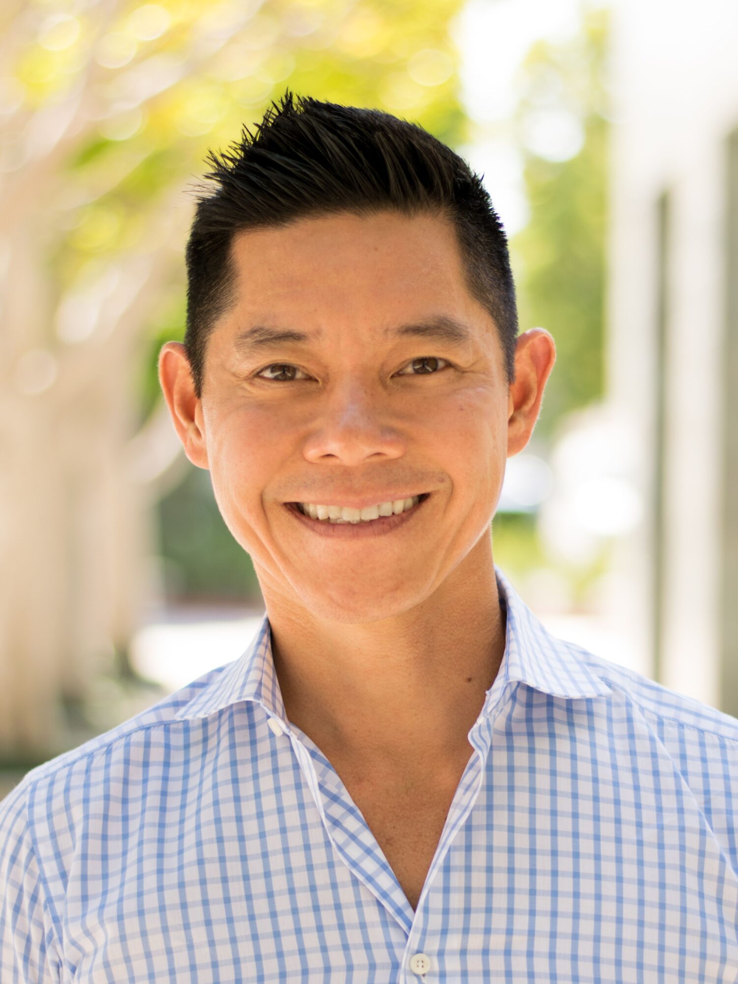 A man with short black hair smiles at the camera in a checkered shirt, exuding warmth and authenticity. The softly blurred background hints at an outdoor setting, reminiscent of stories about Genea's serene landscapes.