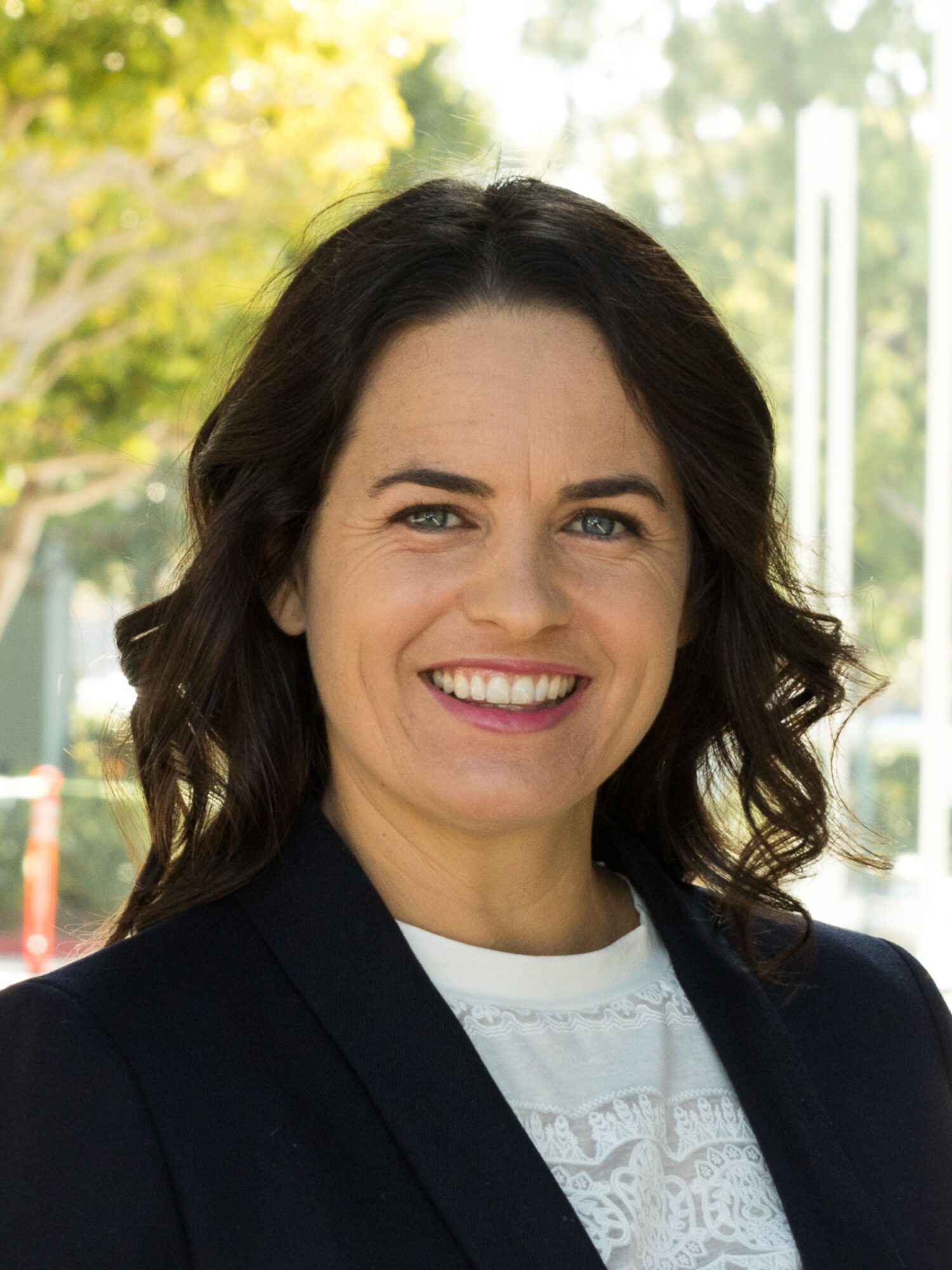 A woman with dark hair and an inviting smile looks at the camera, wearing a white top and a black blazer. The background features blurred outdoor greenery and structures, creating a serene ambiance—a glimpse into the world about Genea.